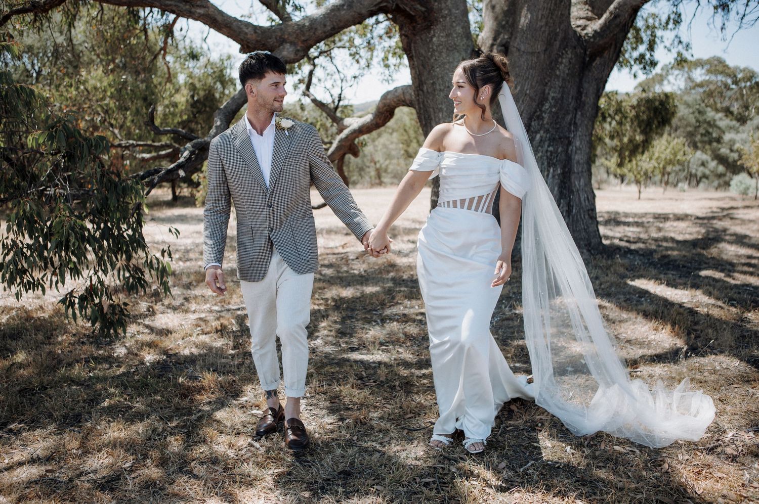Bride and groom hold hands walking under a large tree outdoors, bride wearing white gown with veil.