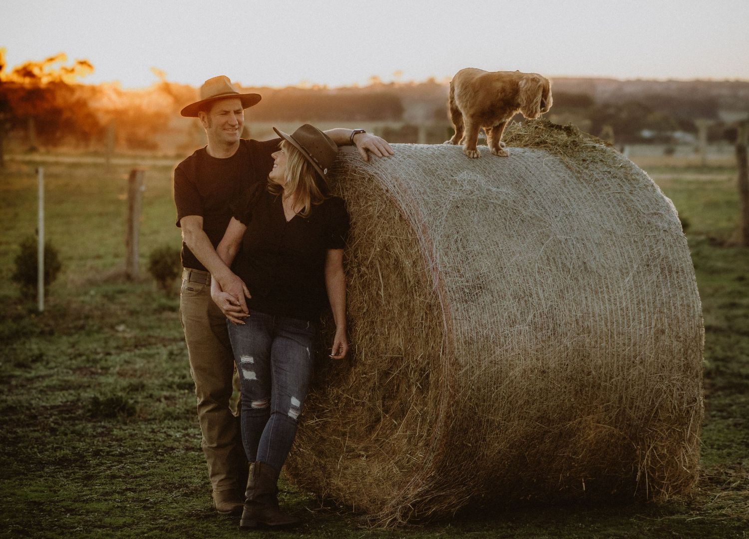Couple leaning against a hay bale with a small animal on top at sunset on a farm.