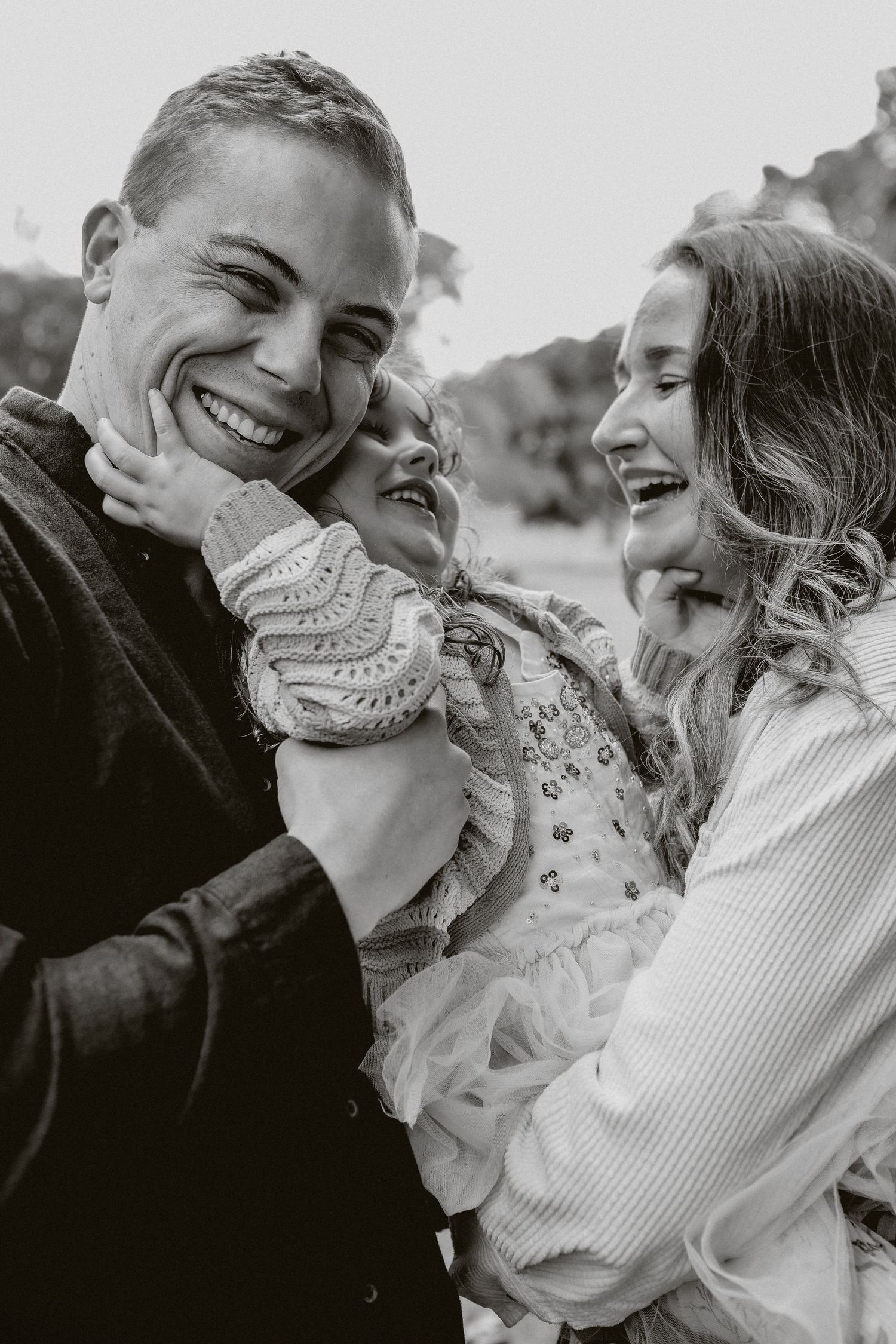 Black and white candid photo of laughing family with child between two parents outdoors.