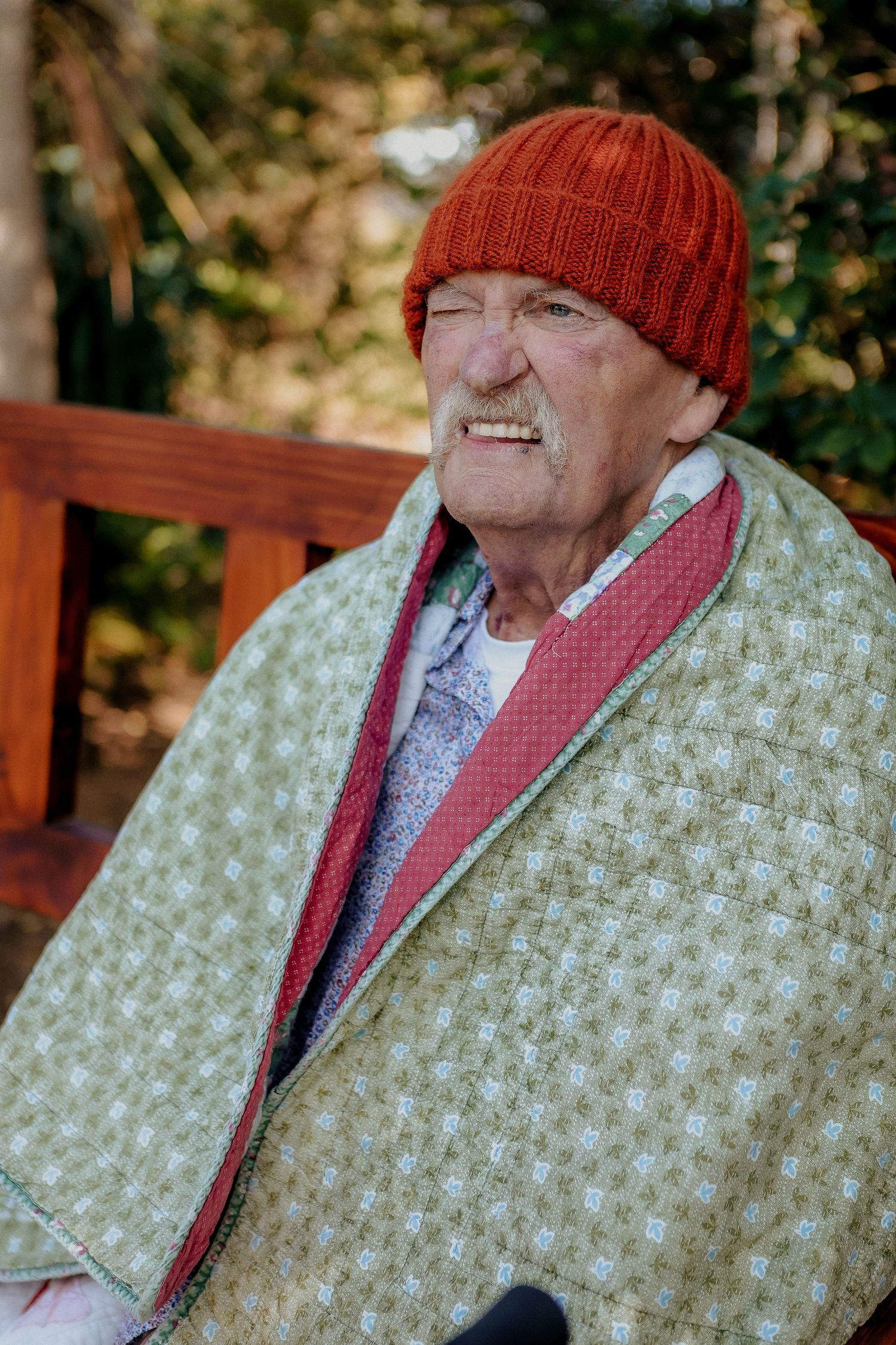 Elderly man wearing an orange knit beanie and green quilt wrap seated on a garden bench.