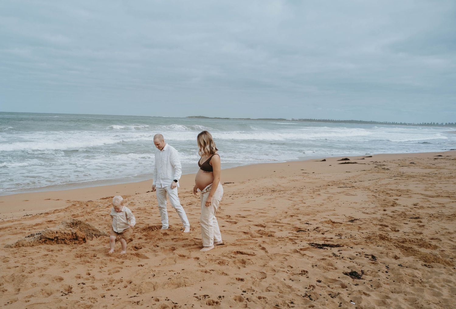 Pregnant woman in bikini walks on sandy beach with partner and toddler, ocean waves in background on cloudy day.