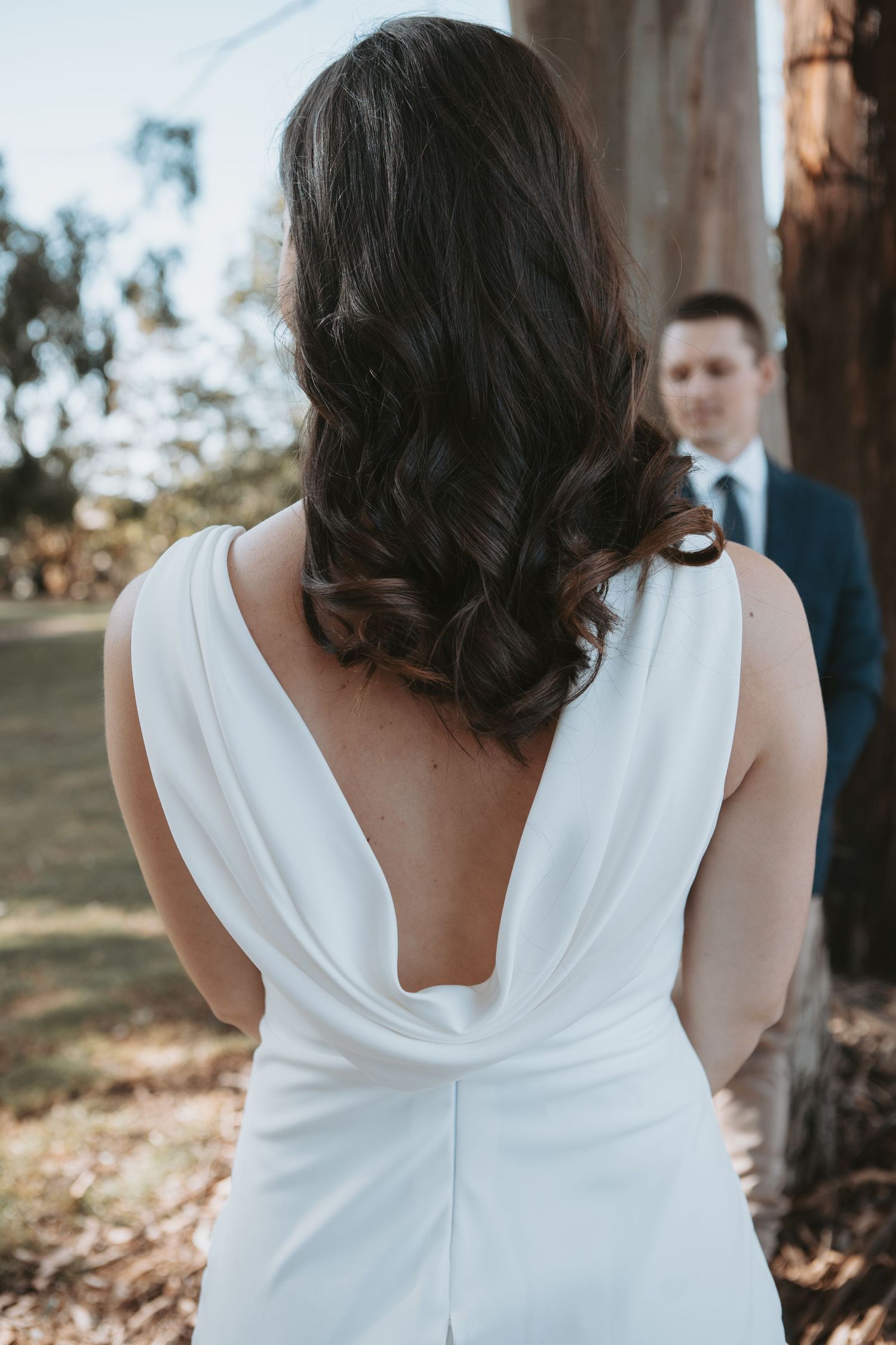 Bride in white open-back wedding gown with dark curly hair, groom in navy suit standing in background outdoors.