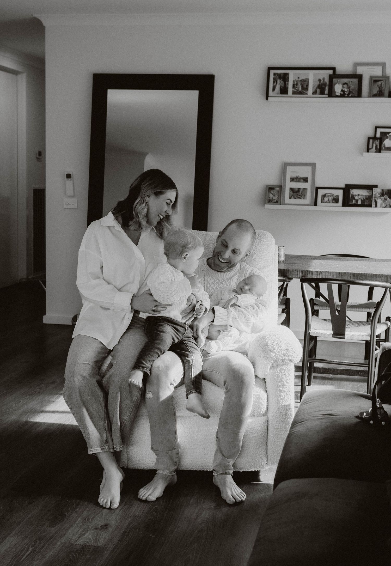 Black and white photo of a family with two young children sitting together in a cozy armchair at home.