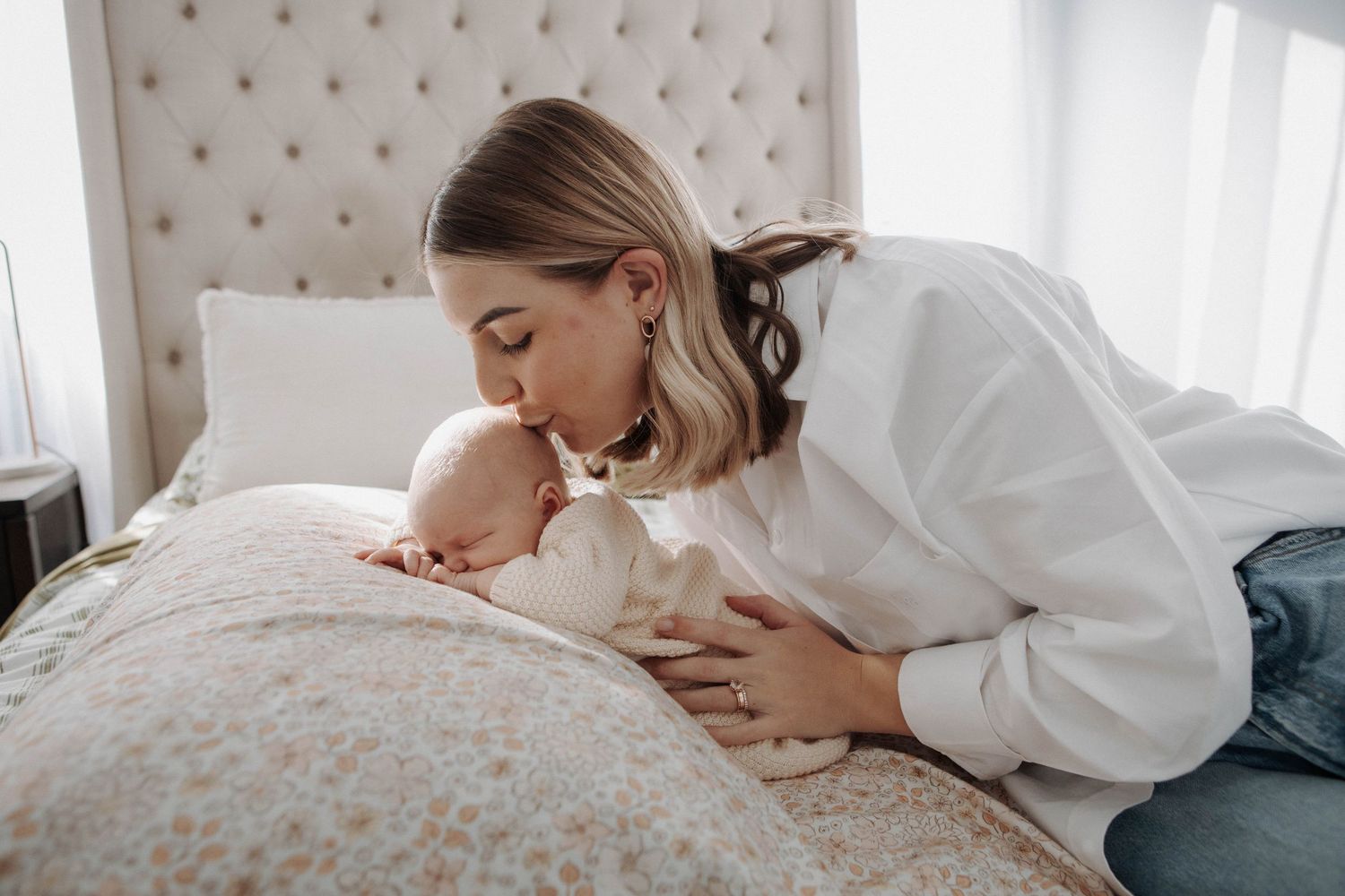 A mother in a white sweater tenderly kisses her newborn baby lying on a floral bedspread.
