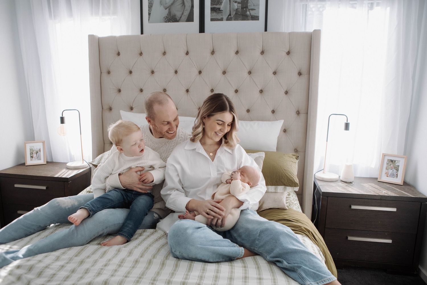 Family of four sitting on a bed, parents smiling with toddler and newborn baby in a cozy bedroom.