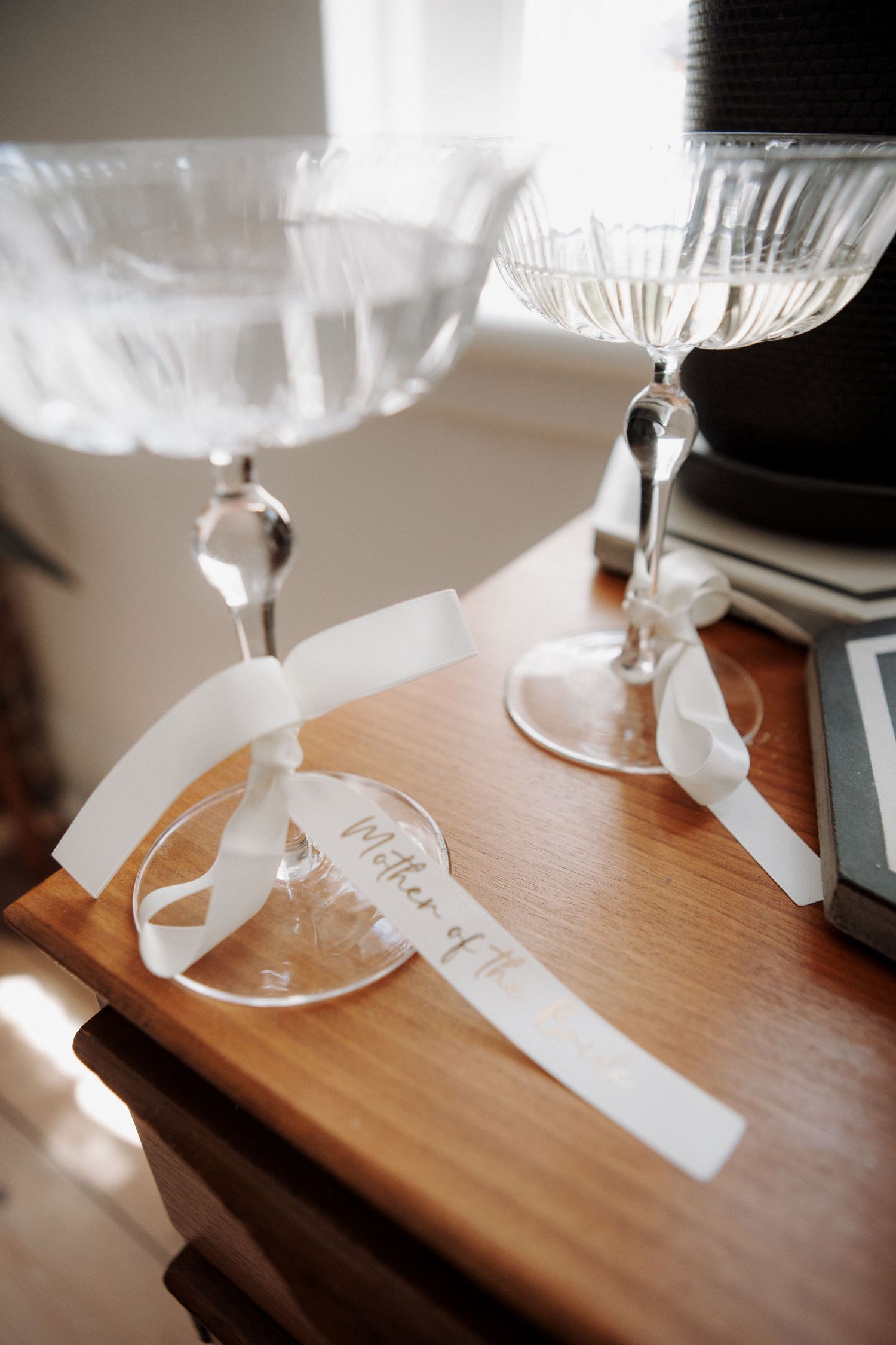 Two crystal coupe glasses with white ribbon name tags labeled Mother of the on a wooden side table.
