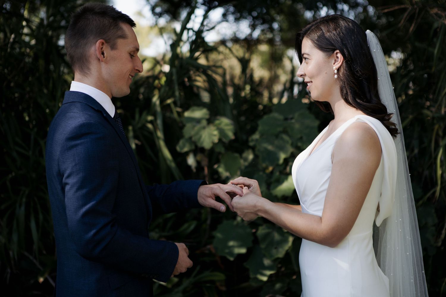 Groom places wedding ring on brides finger during outdoor ceremony surrounded by greenery.