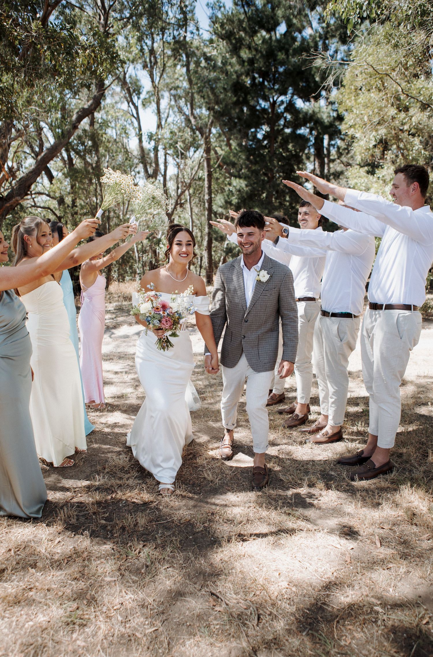 A bride and groom walk hand-in-hand through a guard of honor formed by bridesmaids and groomsmen outdoors.