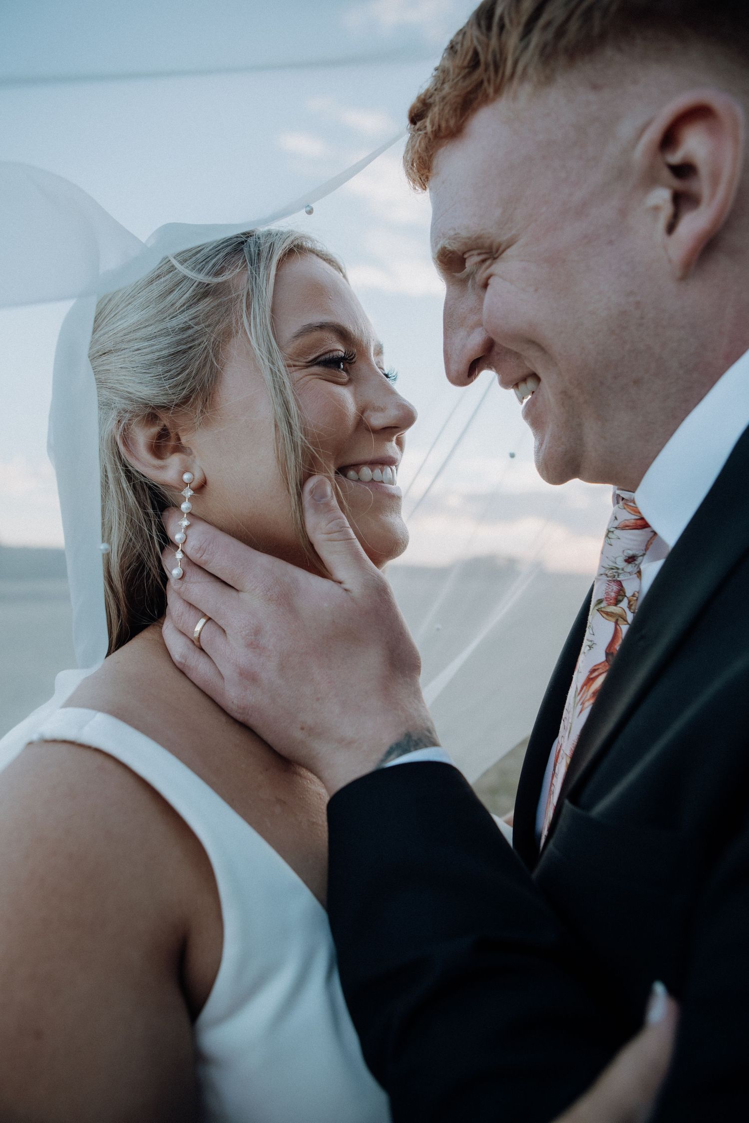 A smiling bride and groom share a tender moment under a veil, with the groom gently cupping her face on their wedding day.