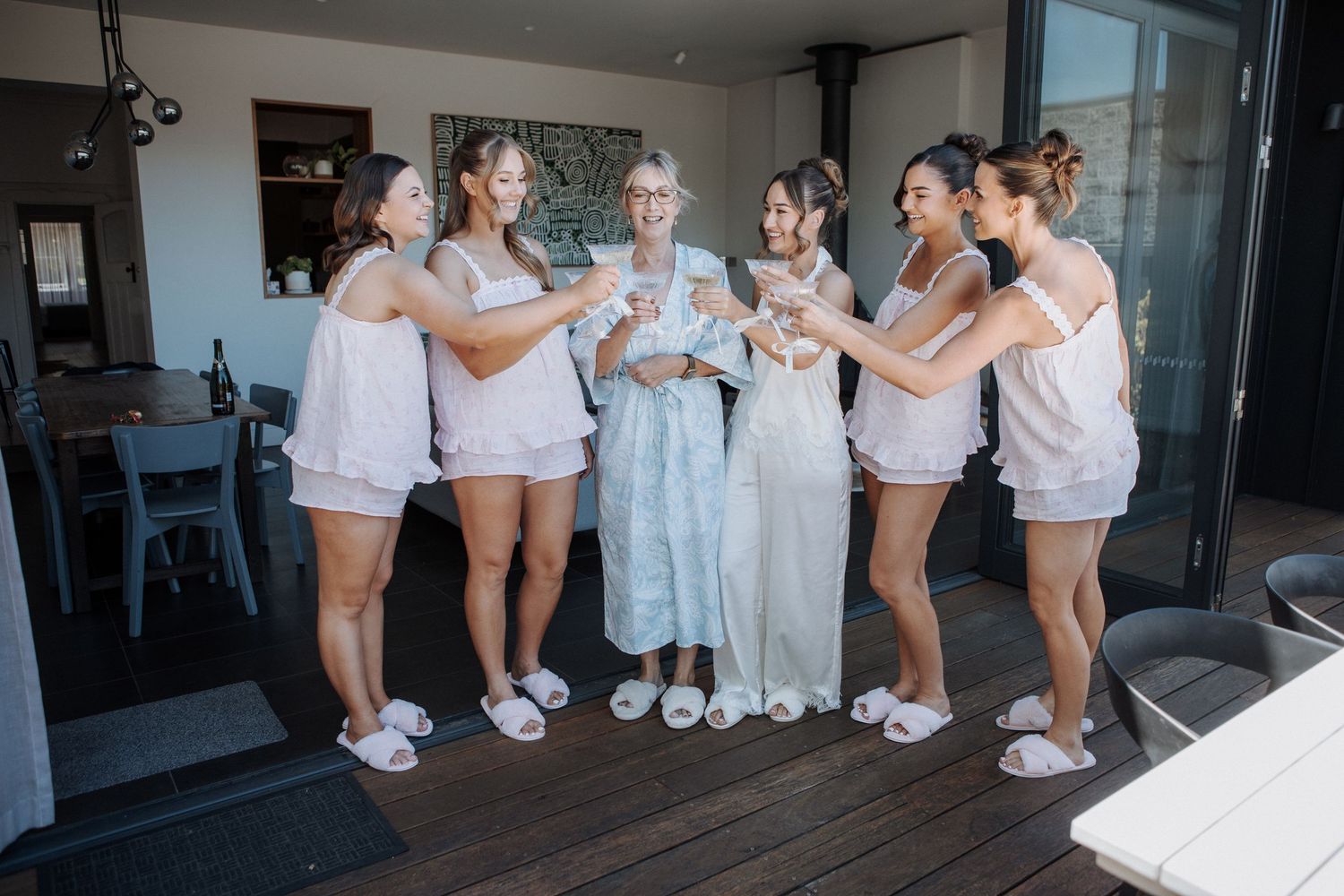 Bride and bridesmaids in white pajamas toasting champagne on deck during wedding morning preparations.