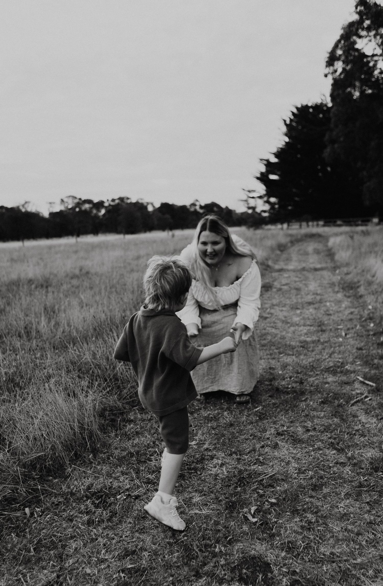 Black and white photo of a mother bending down to greet her young child on a rural grass path.