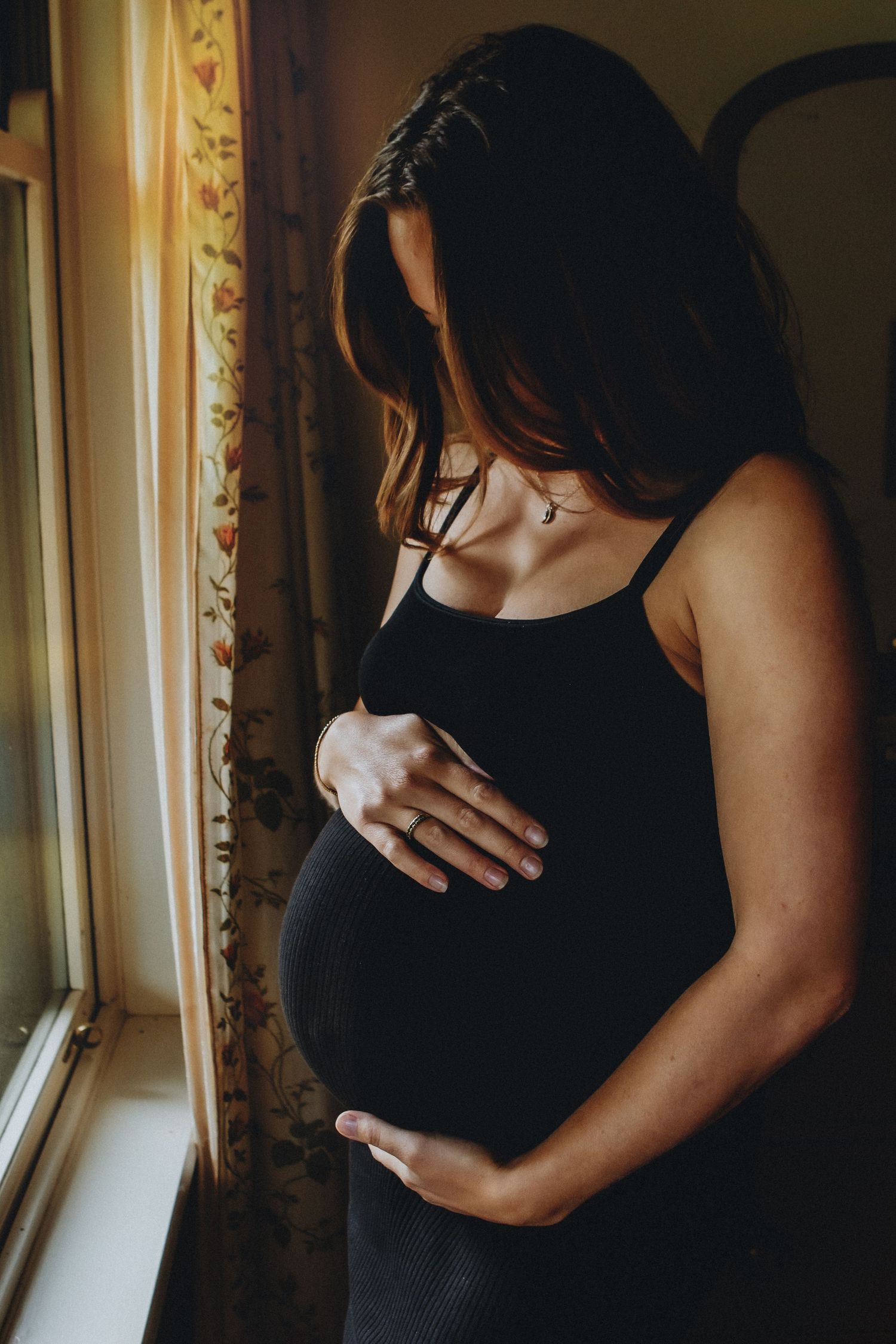 Pregnant woman in black dress gently holding her baby bump while standing by a sunlit window.