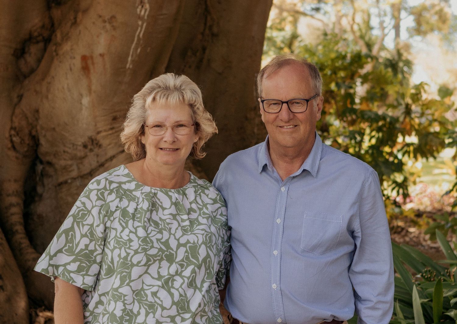 Smiling older couple standing together outdoors in a lush garden beside a large tree trunk at Warrnambool Botanical Gardens.