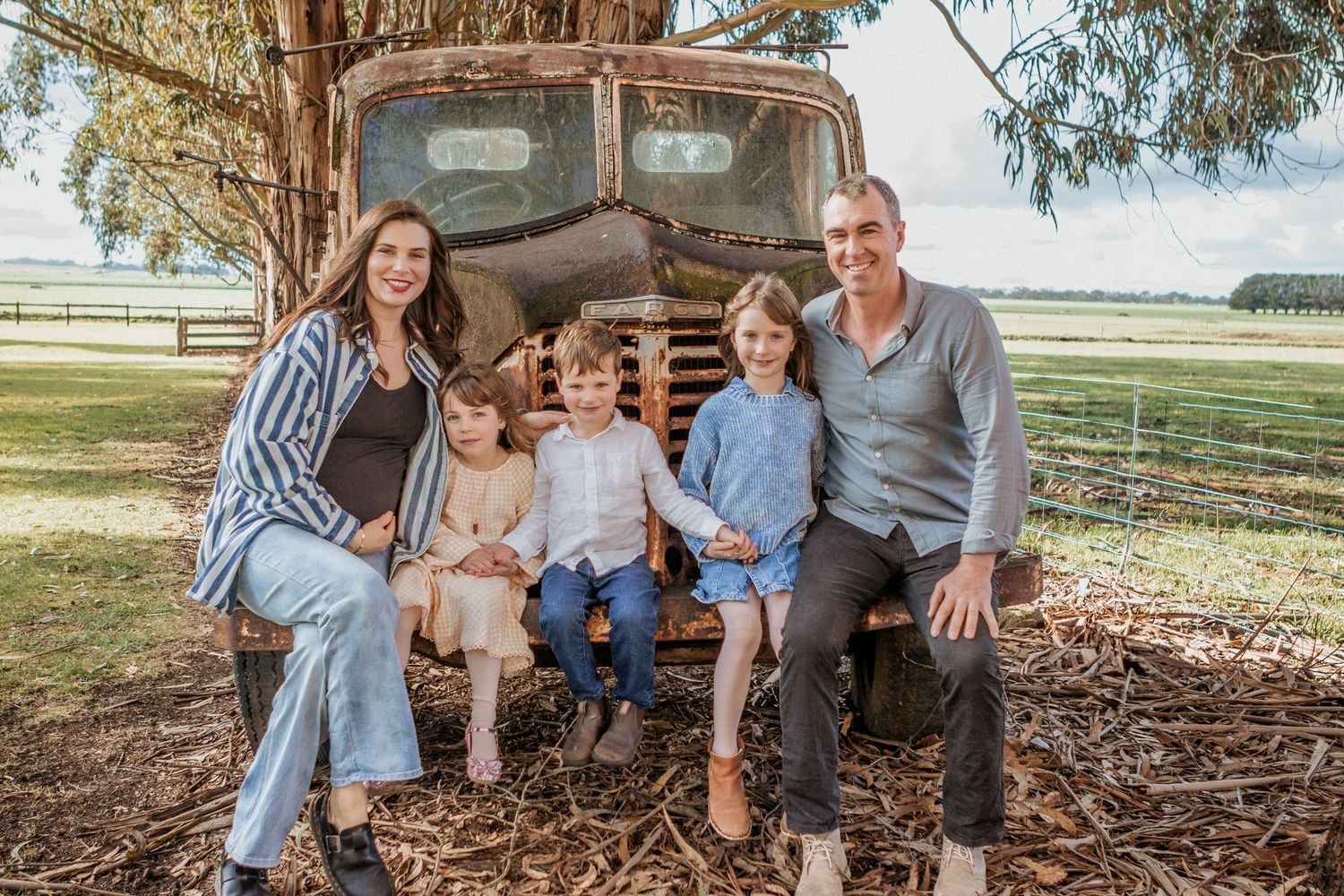 Family of five smiling together on a rustic old truck in a scenic rural farm setting.