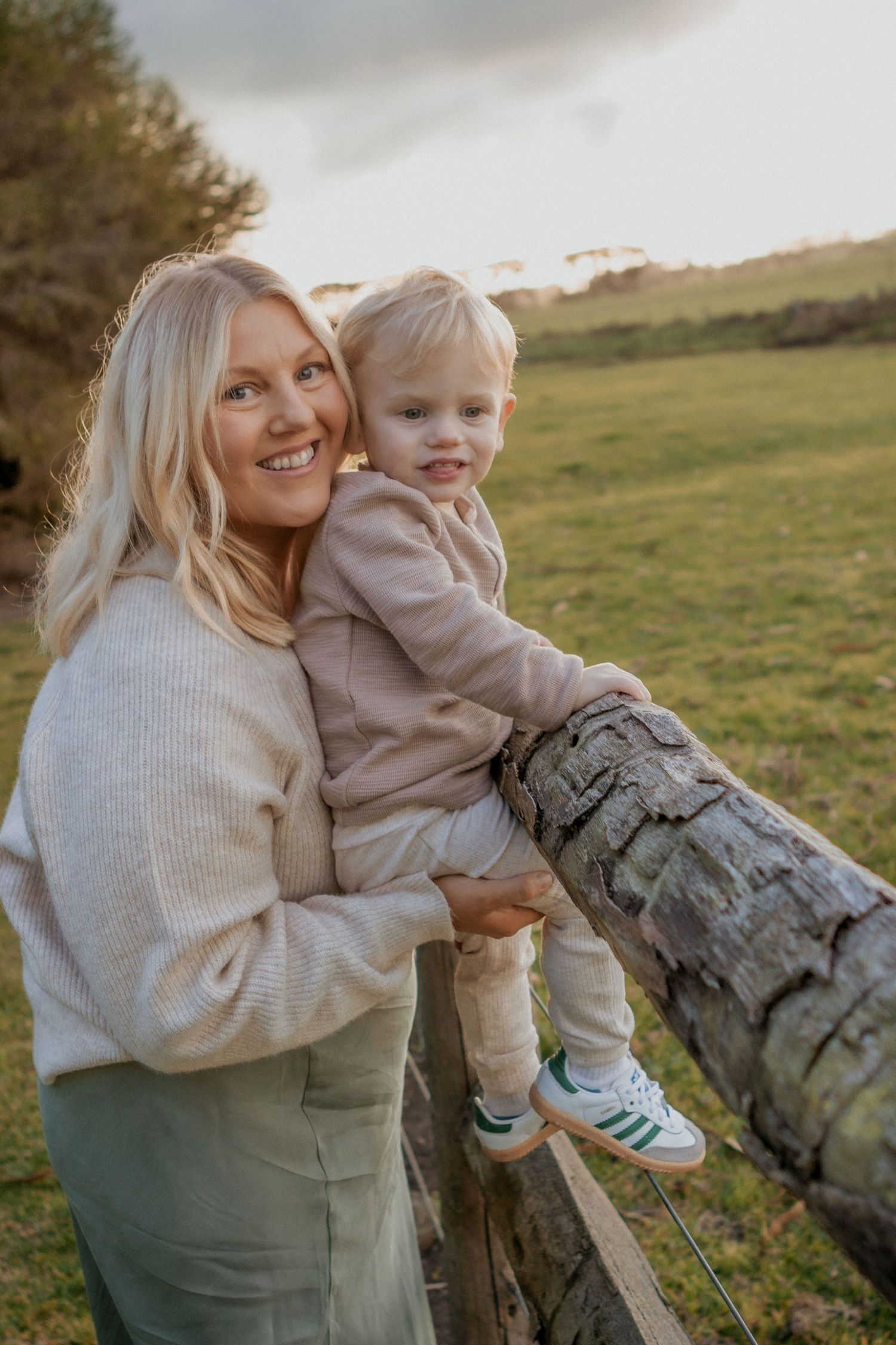 A smiling blonde mother holding her toddler son on a wooden fence in a green field.