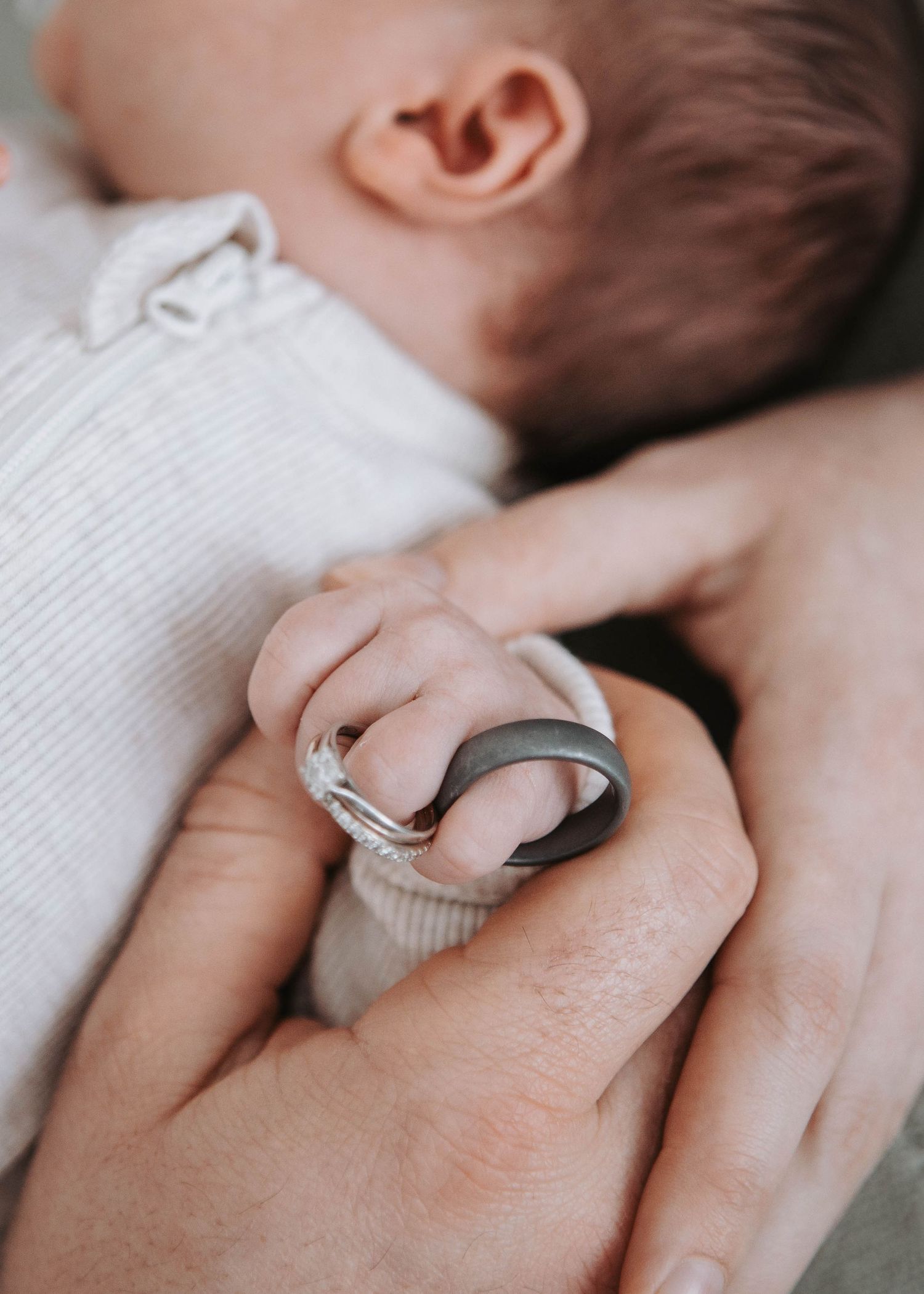 A newborn baby's tiny hand gripping an adult's finger adorned with two rings, symbolizing new parenthood.