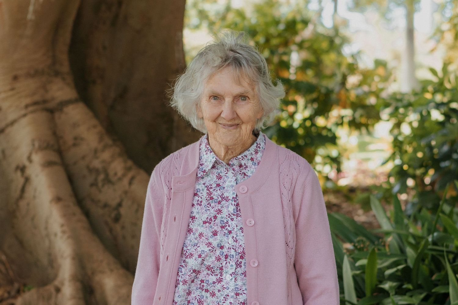 Elderly woman smiling warmly in a pink cardigan standing in front of a large tree in at Warrnambool Botanical Gardens.