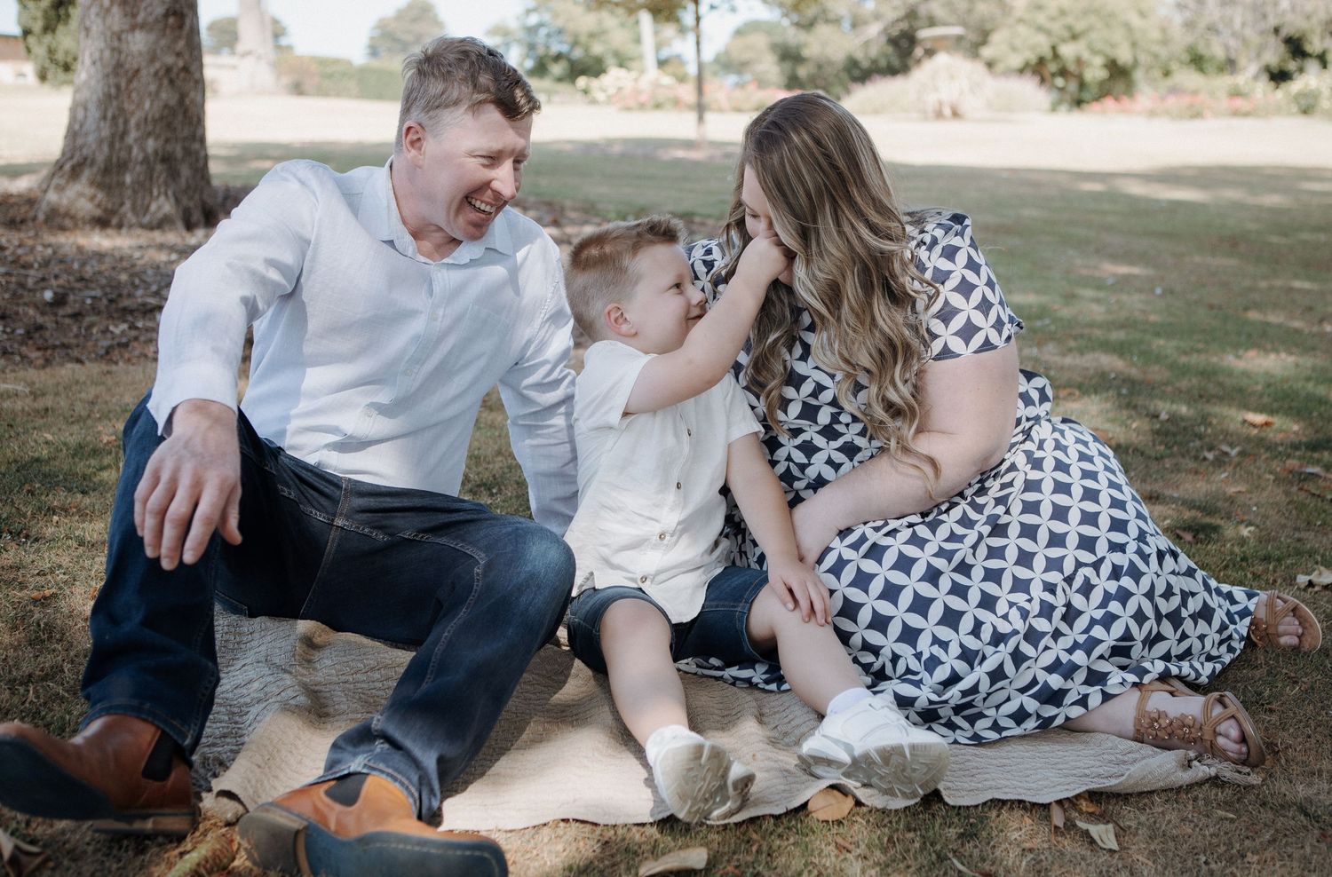 A family of three sits on a blanket in a park, with a toddler boy playfully touching moms face.