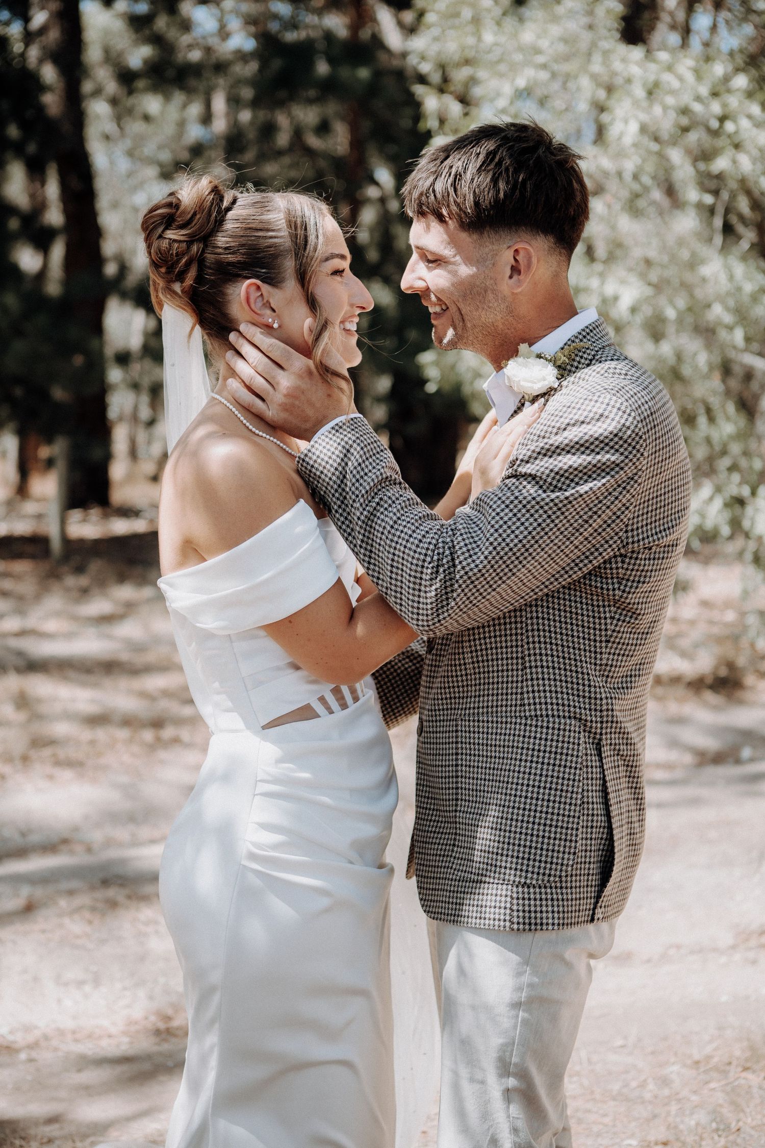 Smiling bride and groom share a tender moment in a sunlit forest, with groom cradling brides face in his hands.