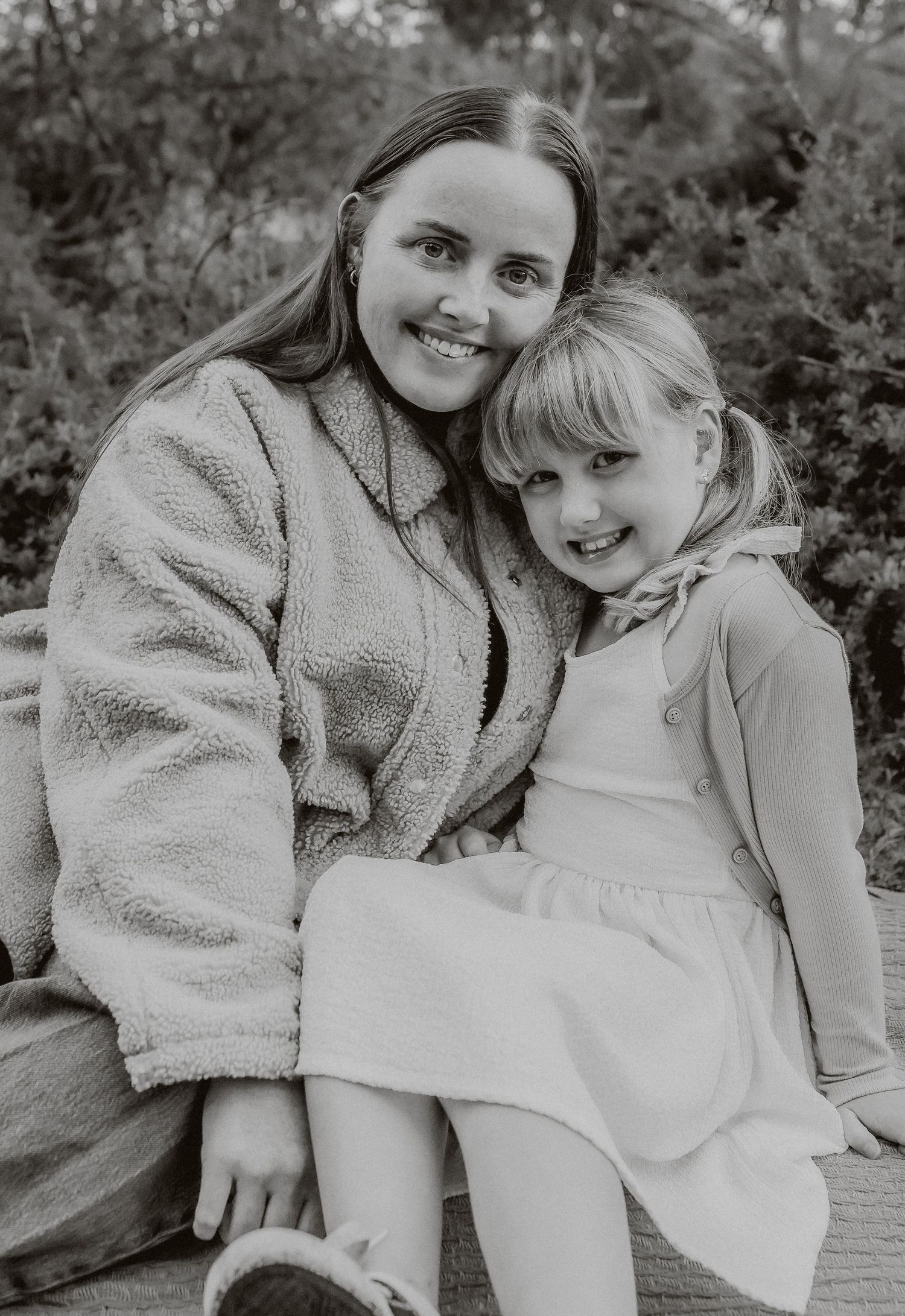 Black and white portrait of a smiling mother and young blonde daughter sitting together outdoors.