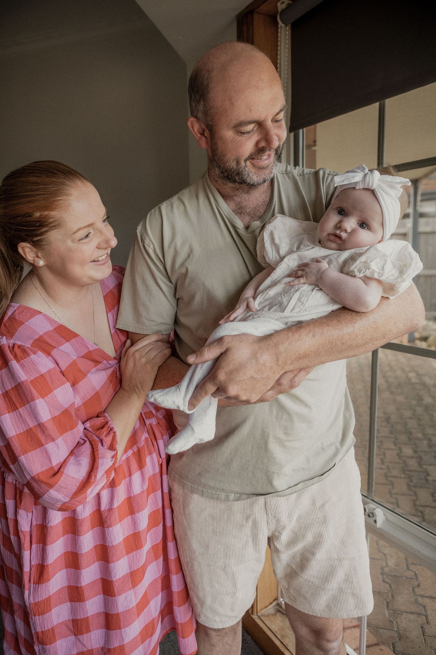 Proud parents in pink dress and beige outfit lovingly holding newborn baby girl in white outfit.