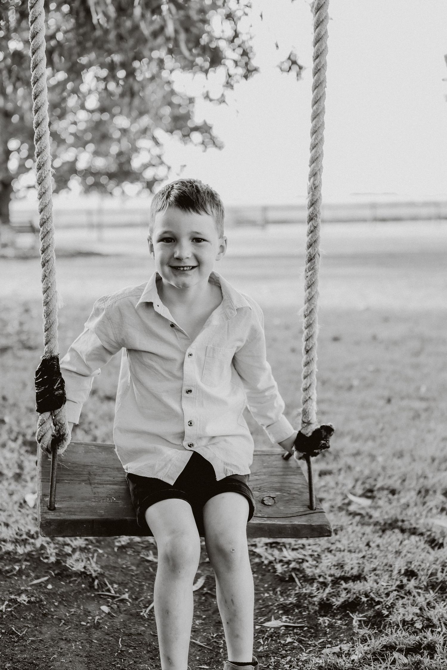 A smiling boy in a white shirt sits on a wooden rope swing outdoors in a black and white photo.