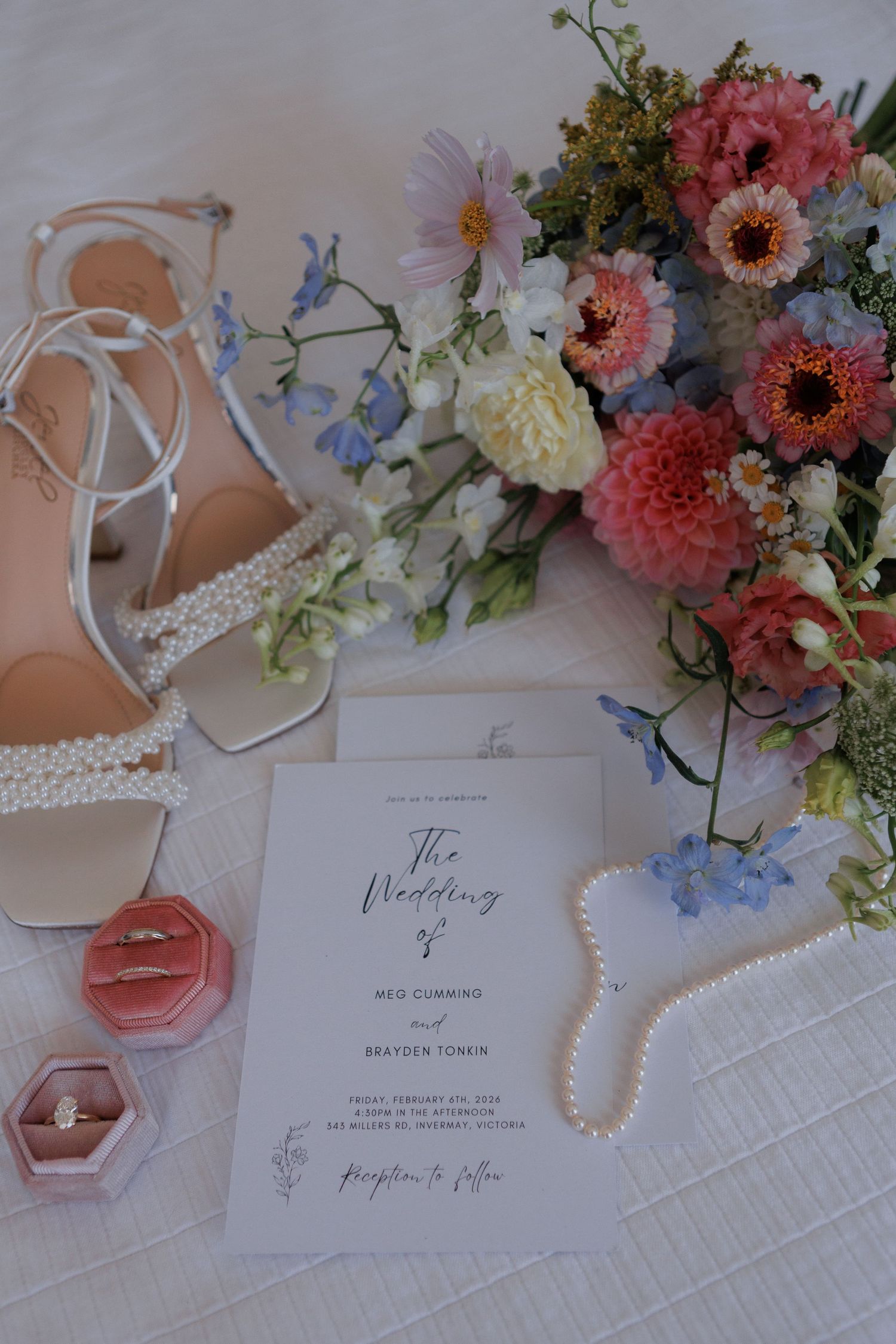 Elegant wedding flat lay with pearl heels, floral bouquet, rings in velvet box, pearl necklace, and wedding program on white surface.