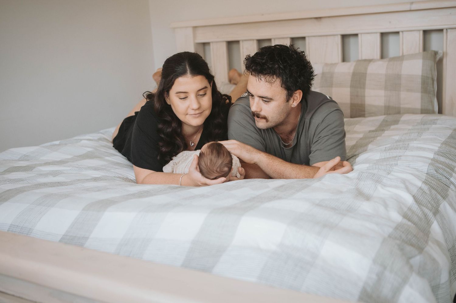New parents lying on bed gazing lovingly at their newborn baby wrapped in white blanket.