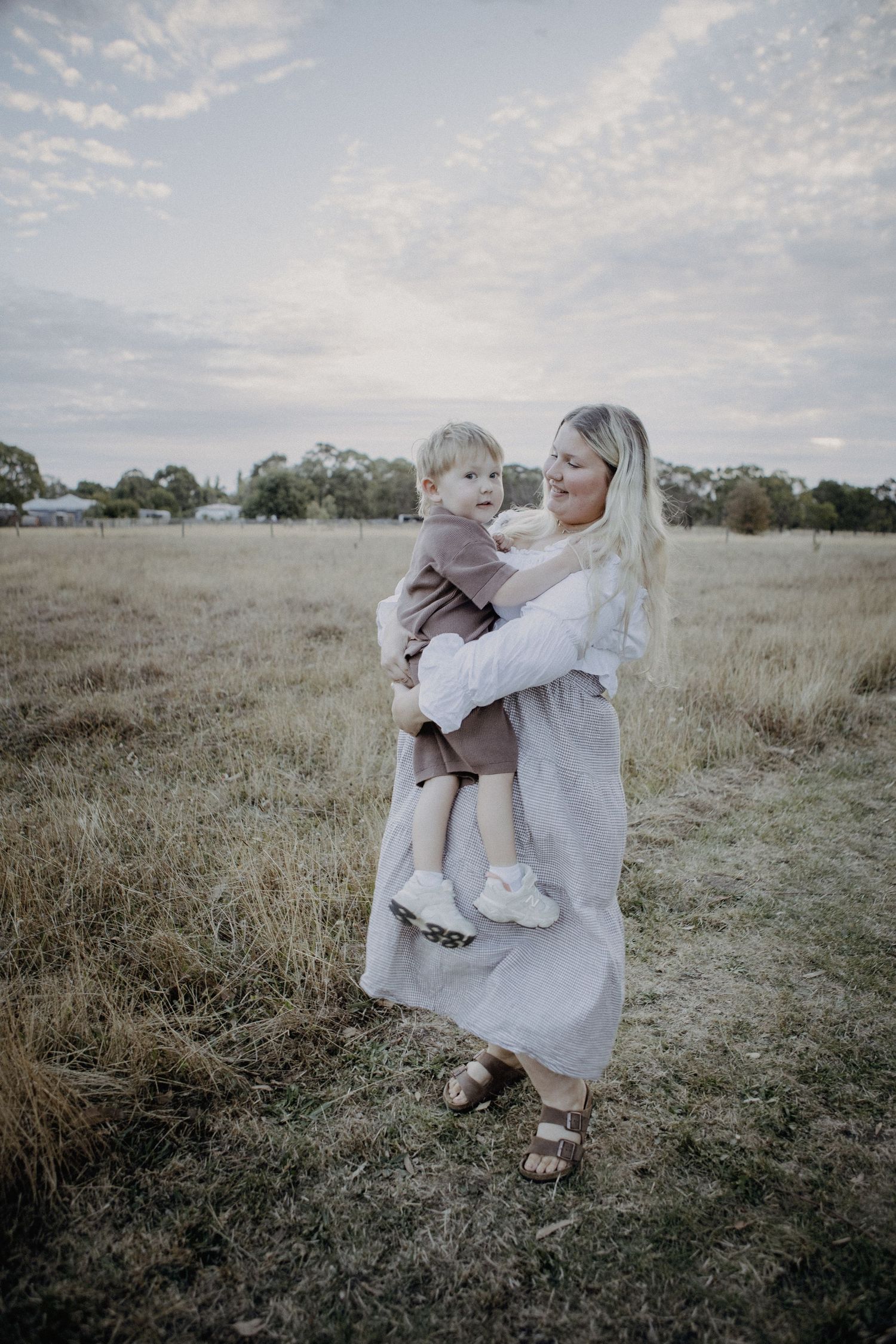 A smiling blonde mother holds her young child in an open field under a cloudy sky.