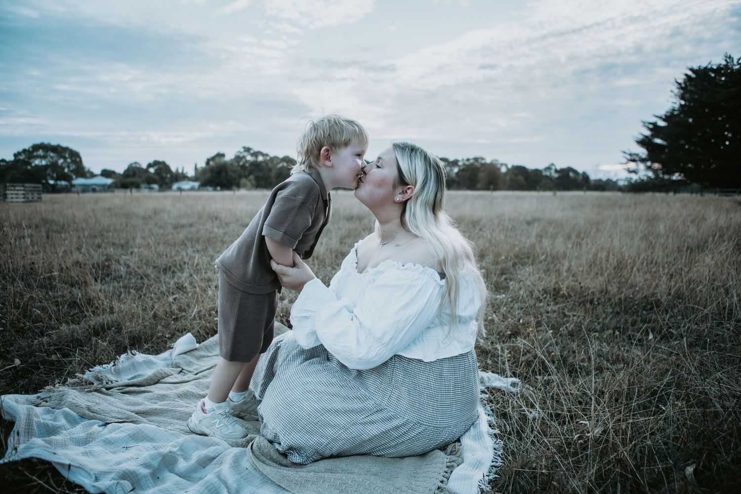 A toddler gives his seated mother a sweet kiss on a blanket in a golden field.