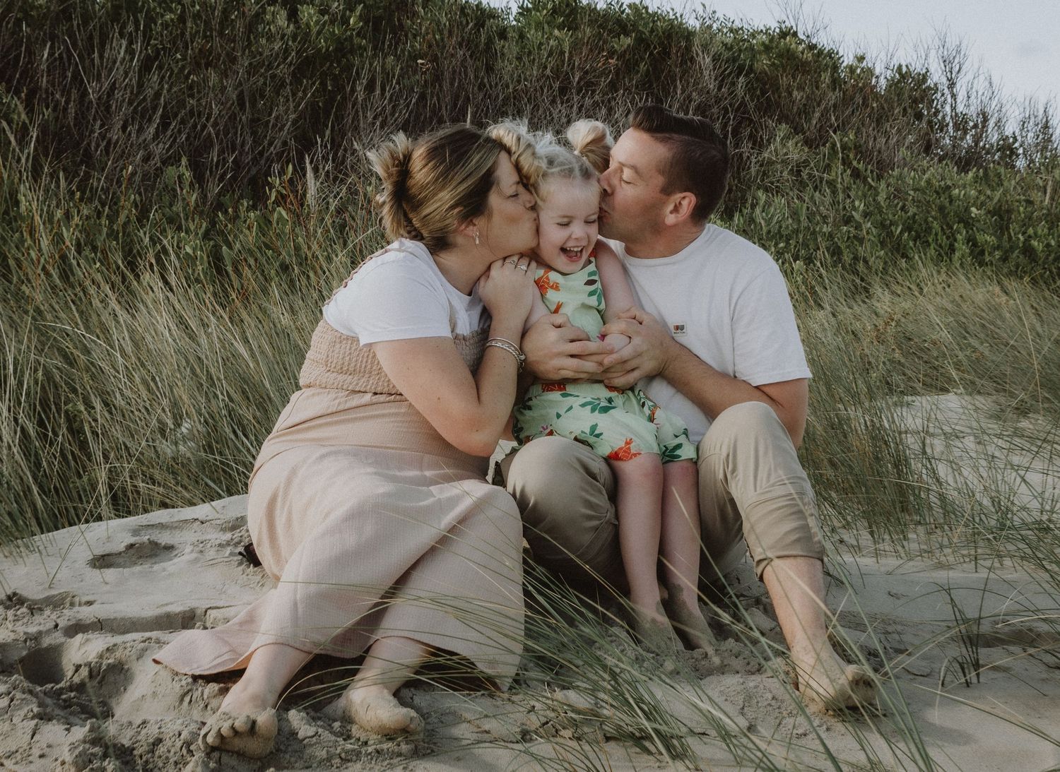 Happy family of three with parents kissing toddler daughter on beach sand dunes.