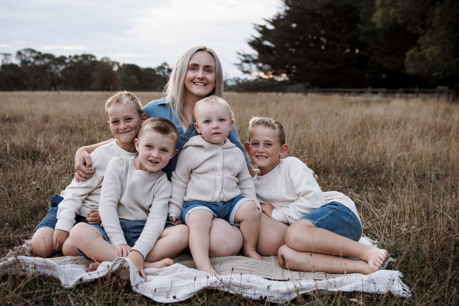 Smiling blonde mother sits on blanket in golden field in Warrnambool with her four young children.