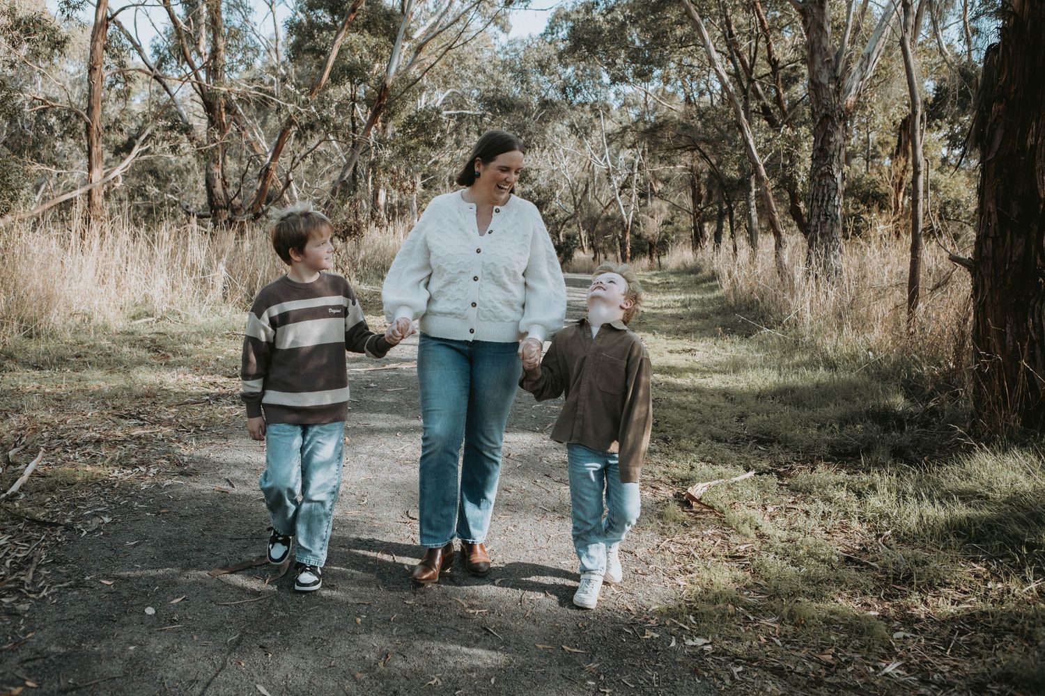 A mother walks hand-in-hand with two children on a forest path surrounded by tall trees.