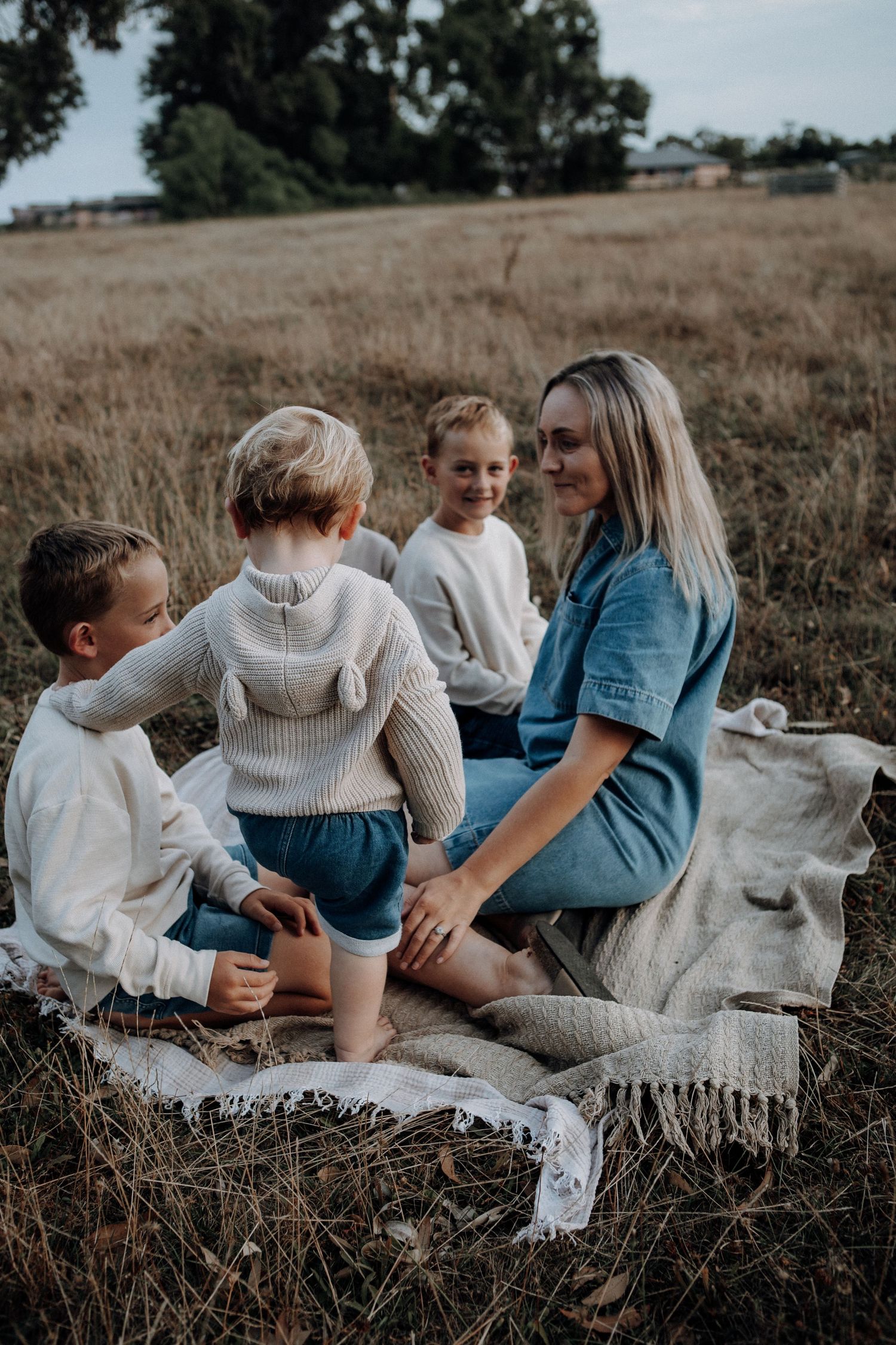 Mother in denim outfit sits on blanket in dry grass field laughing with her three young boys.