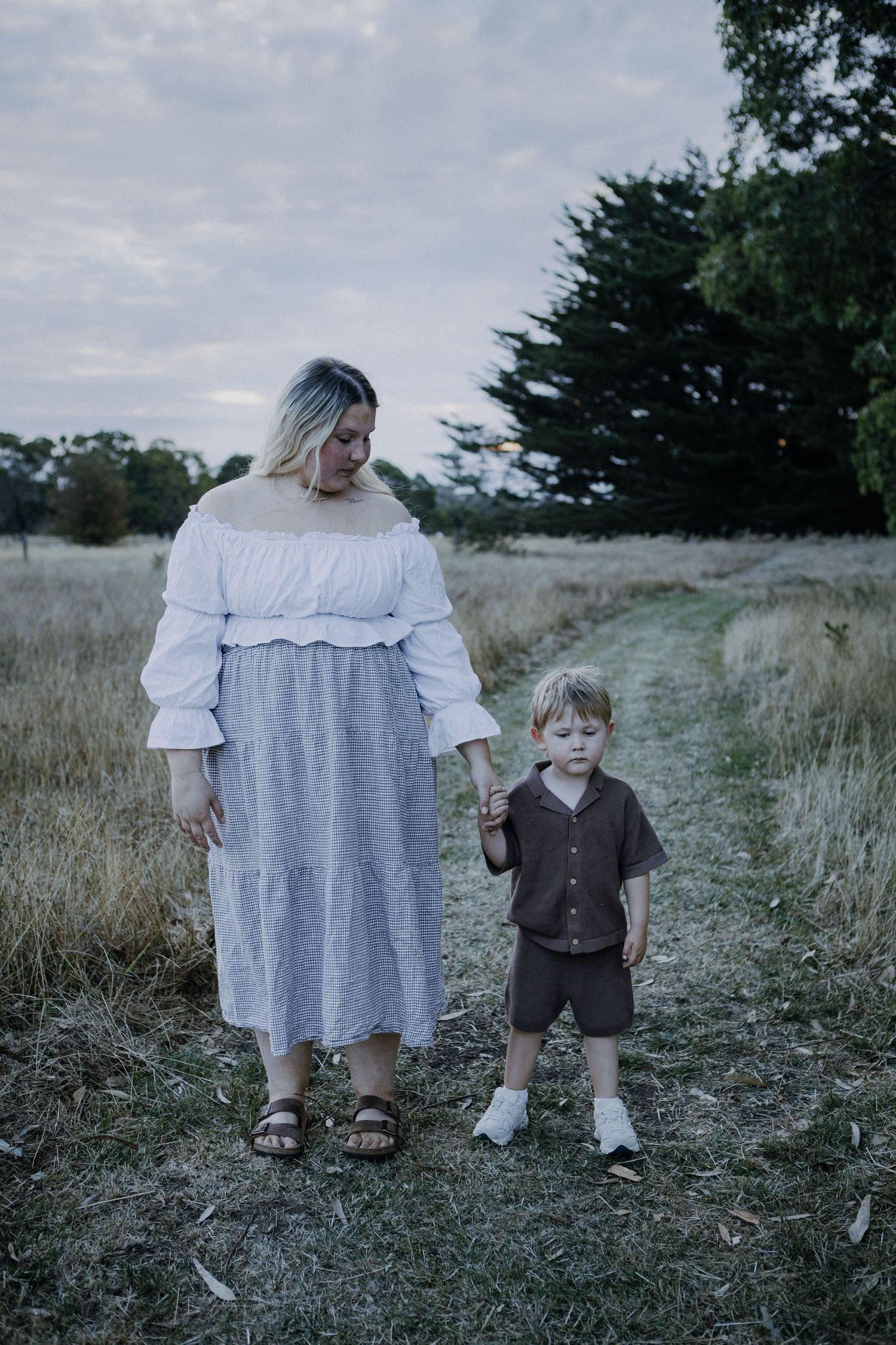 A mother in a white off-shoulder top holds her toddler sons hand on a rural dirt path.
