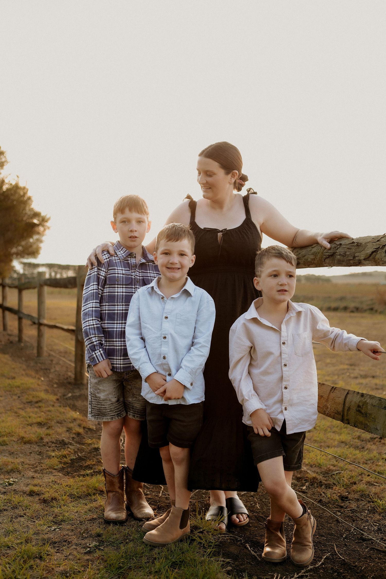 Mother standing with three young sons at golden hour near rustic wooden farm fence.
