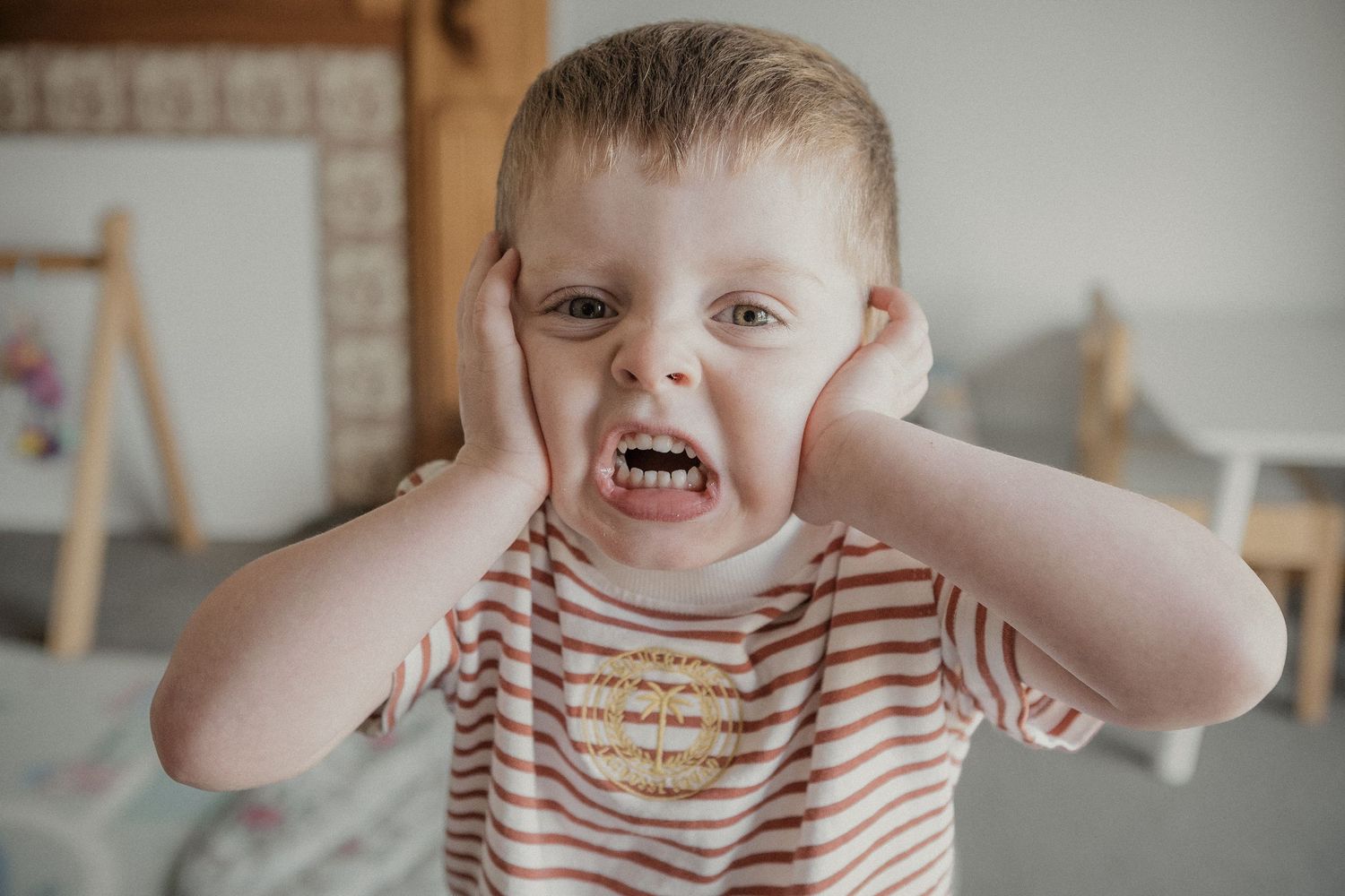 Expressive young toddler boy covering ears making funny face wearing striped shirt indoors.