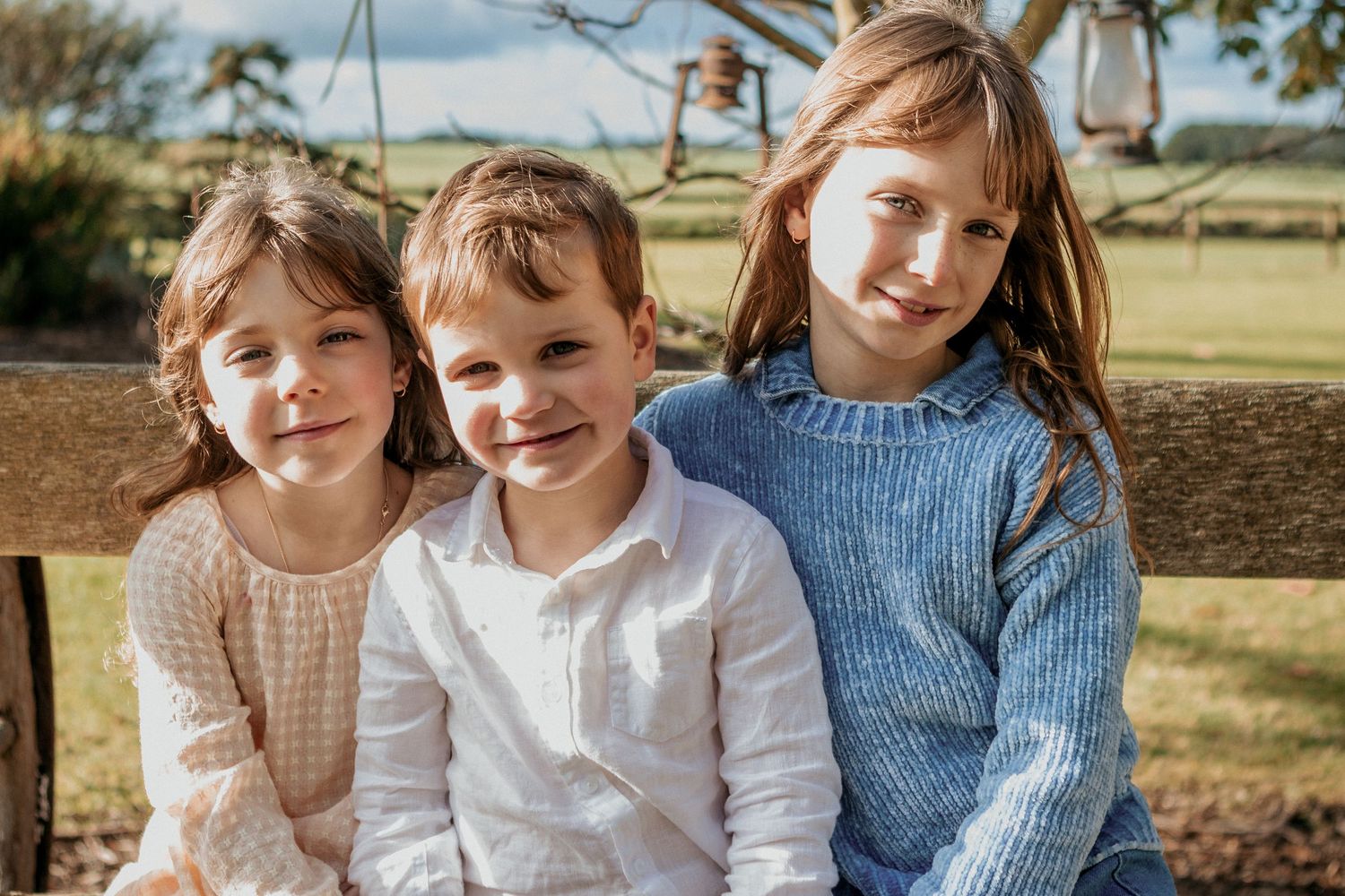 Three young siblings sitting together outdoors smiling in a bright countryside setting.