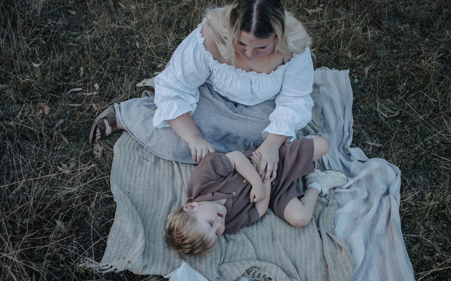 A mother sits on a blanket in a grassy field cradling her sleepy toddler child.