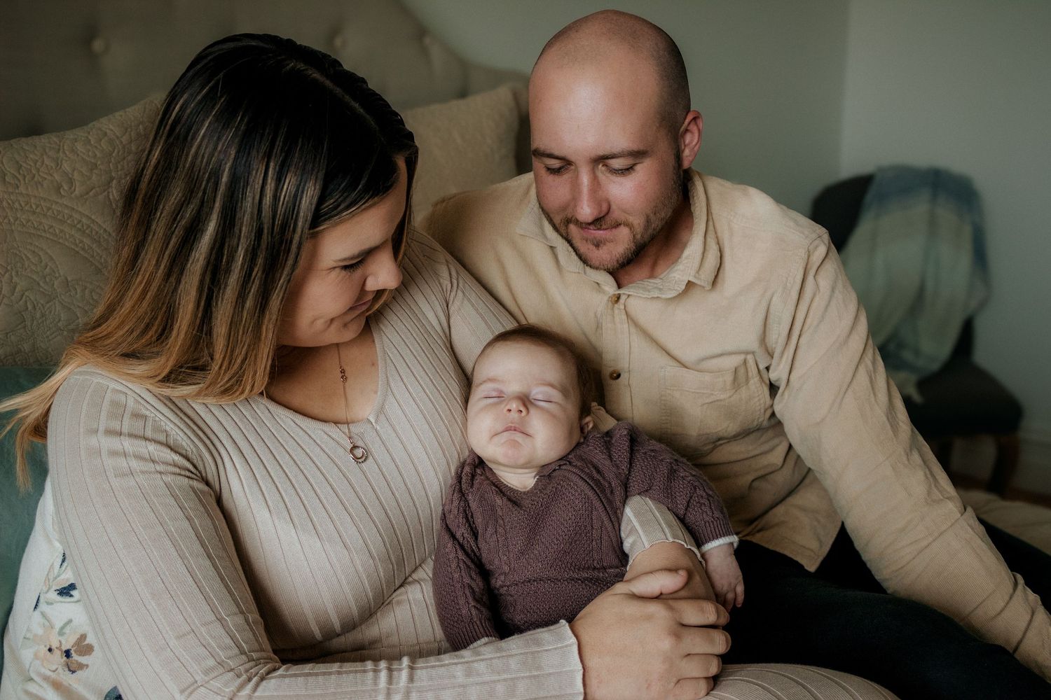 New parents smiling together while cradling sleeping newborn baby in soft bedroom light.