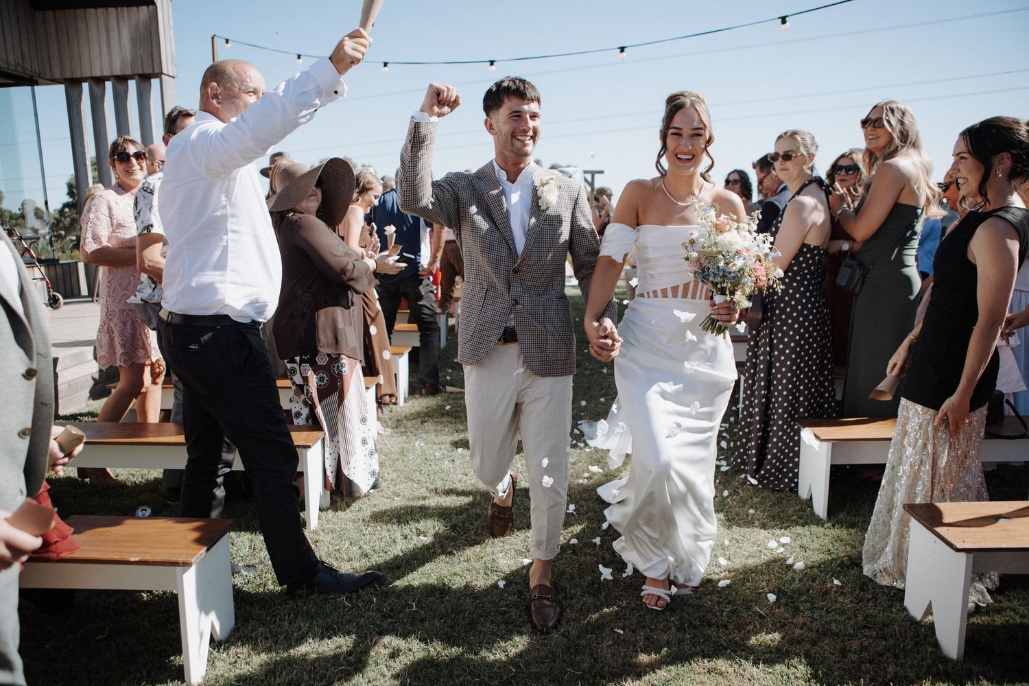 Joyful newlyweds walk down the aisle as guests celebrate at an outdoor wedding ceremony.
