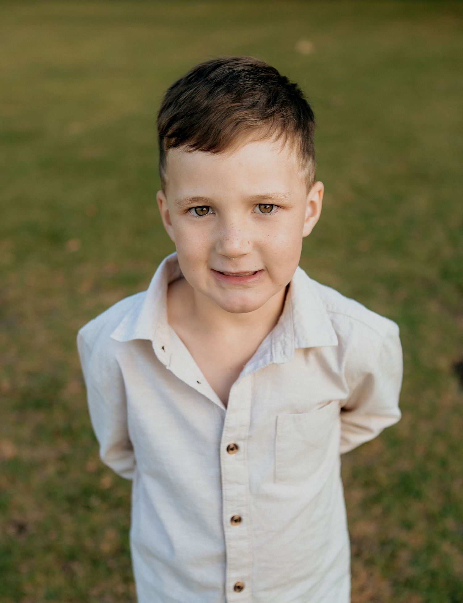 Portrait of smiling young boy in white linen shirt standing on green grass outdoors.