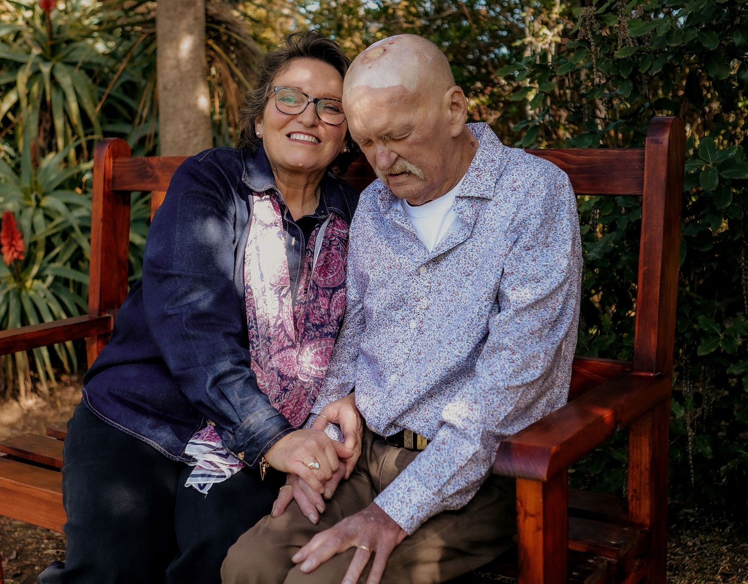 Elderly couple sitting closely together on a red wooden bench in a garden setting.