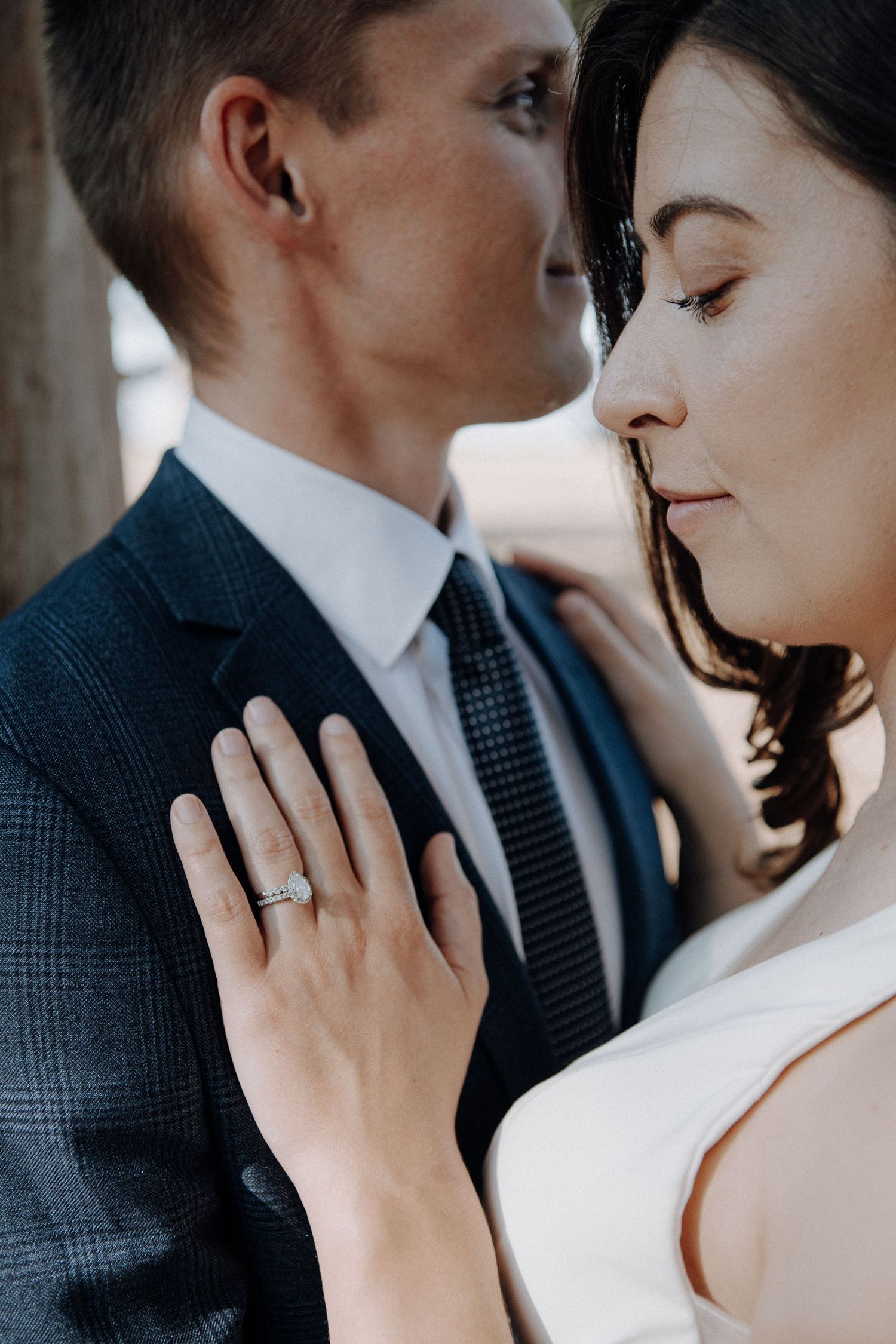 Bride and groom share intimate moment, woman wearing diamond engagement ring resting hand on grooms navy suit jacket.