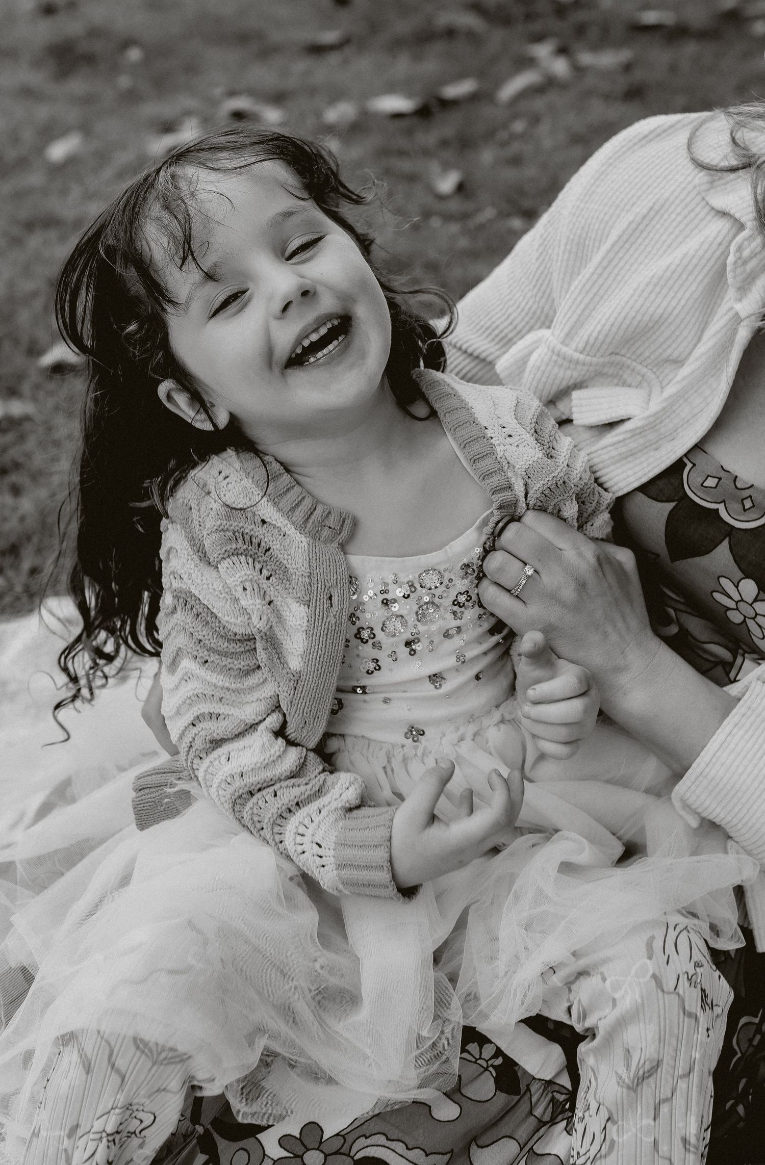 A laughing young girl in a tulle dress and cardigan is held by an adult in a black and white portrait.