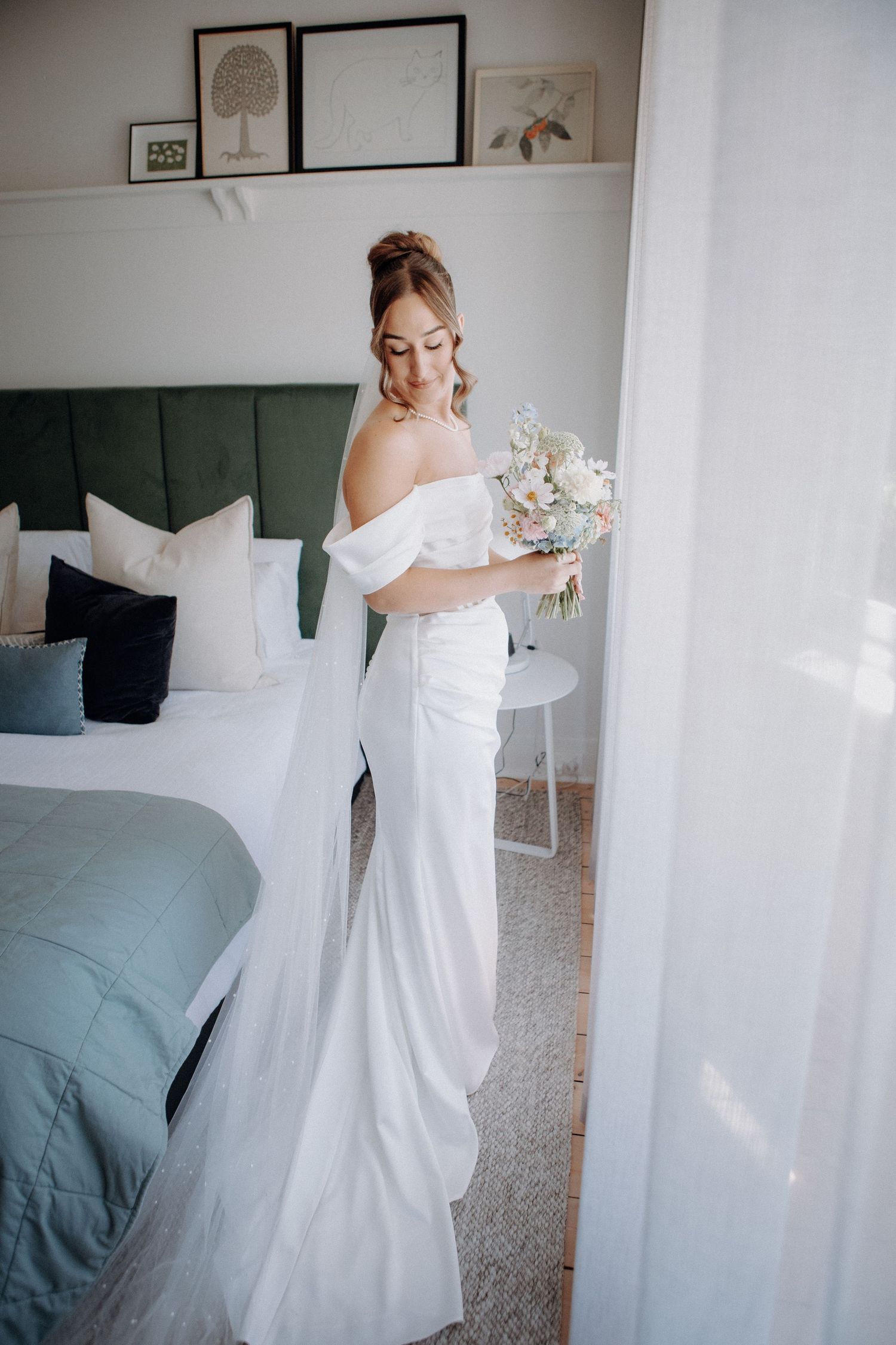 Bride in off-shoulder white gown holding a bouquet in a bright bedroom with green headboard.