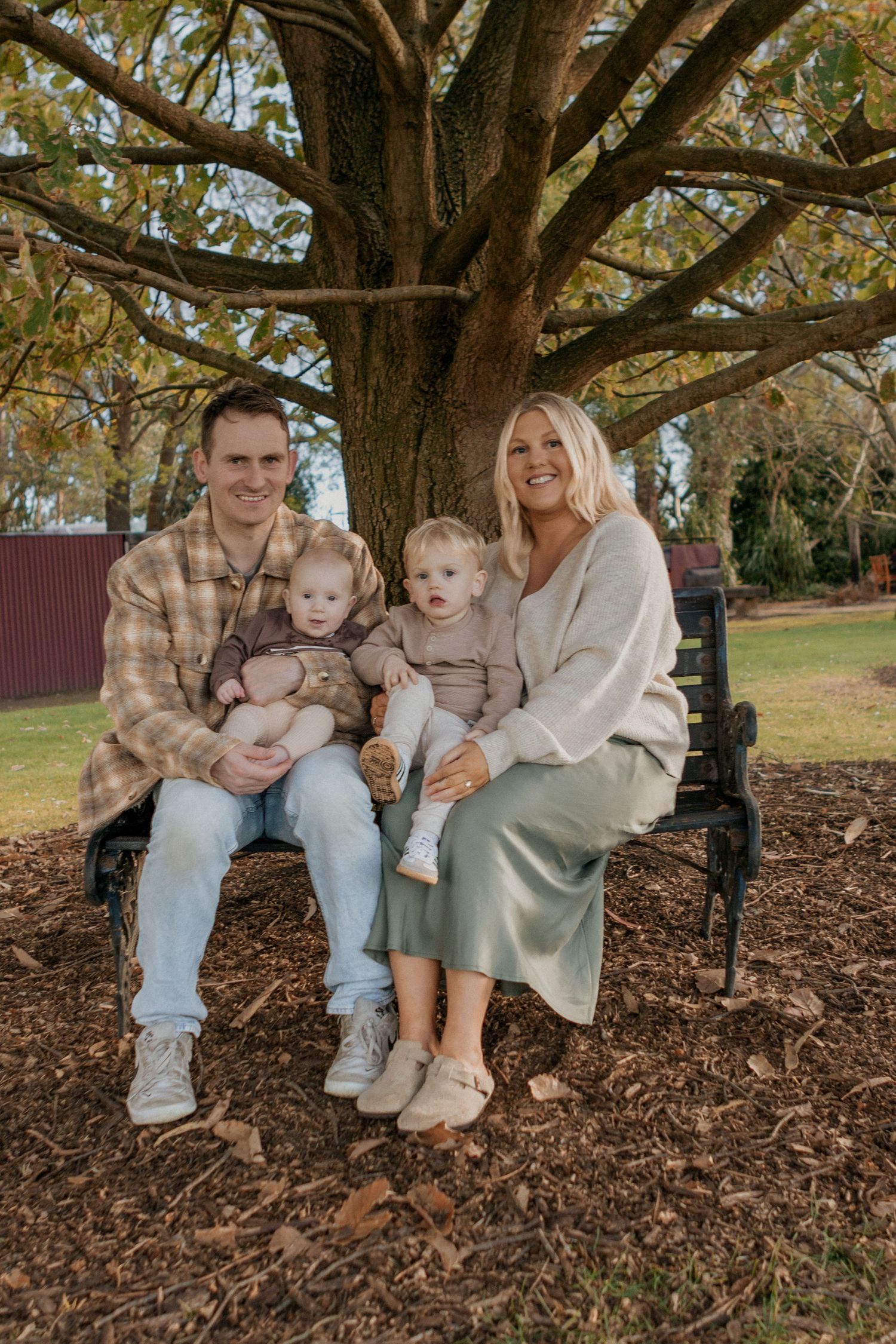 A family of four sitting on a park bench under a large autumn tree, smiling warmly.