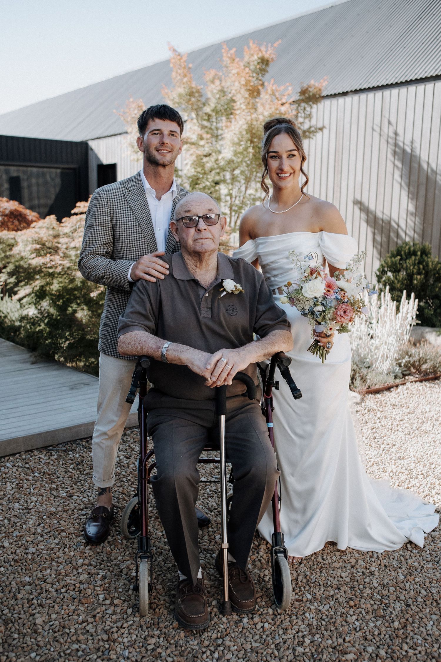 Newlywed couple poses with elderly man in wheelchair outside modern wedding venue.