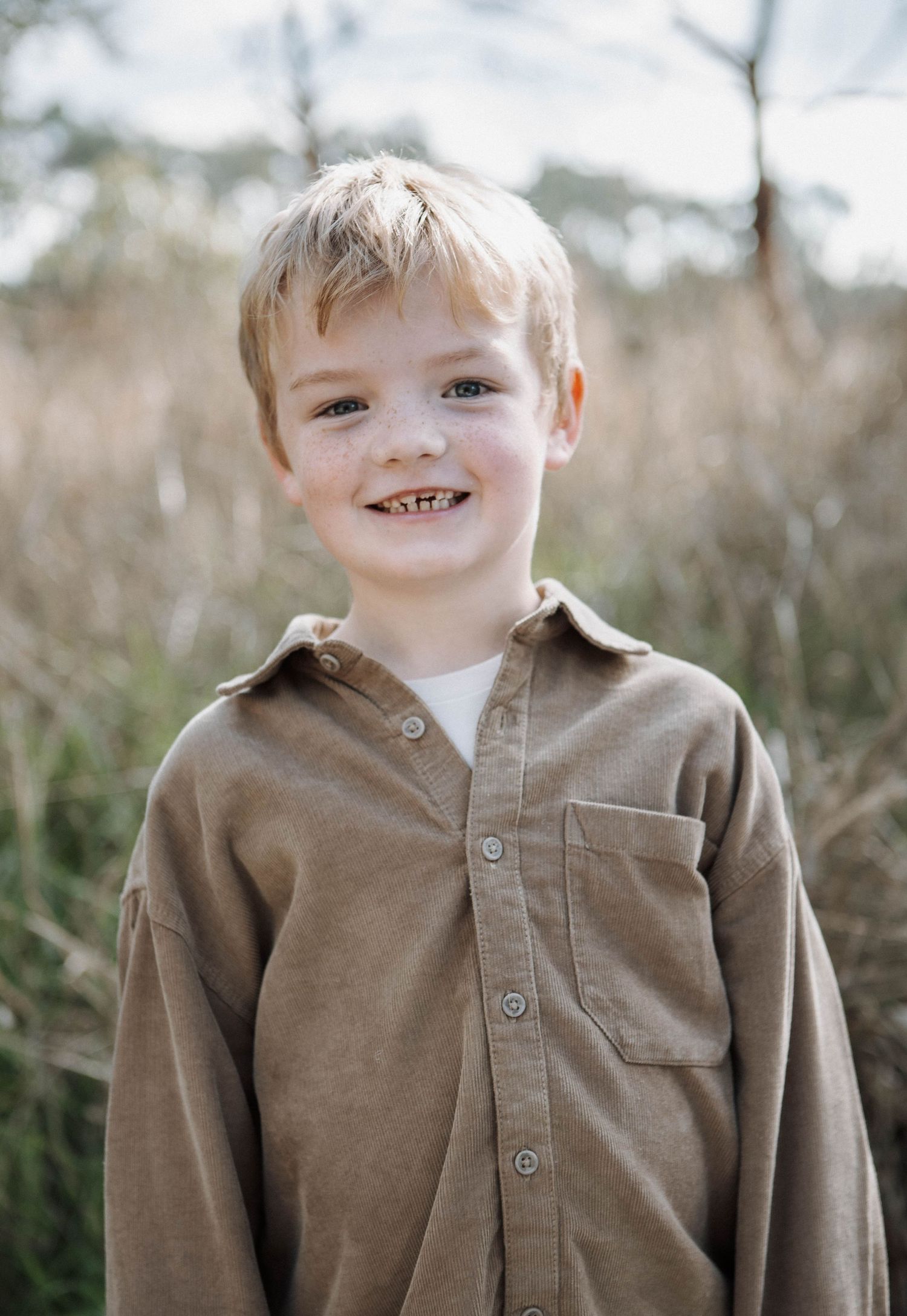 Smiling young boy with blonde hair wearing a brown button-up shirt outdoors in a natural field setting.