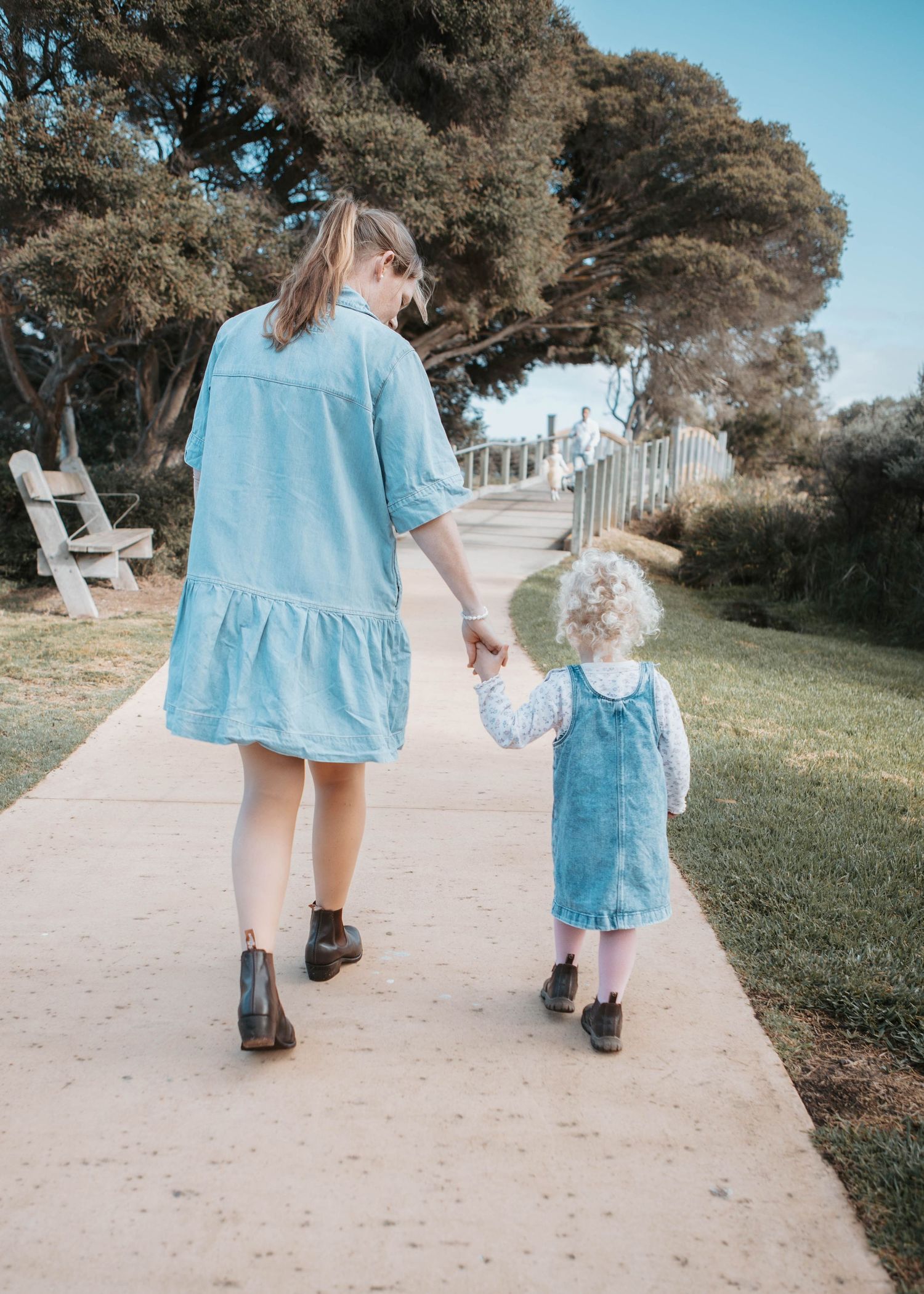 A mother and young daughter in matching denim outfits walk hand in hand along a sunny park path surrounded by green trees.